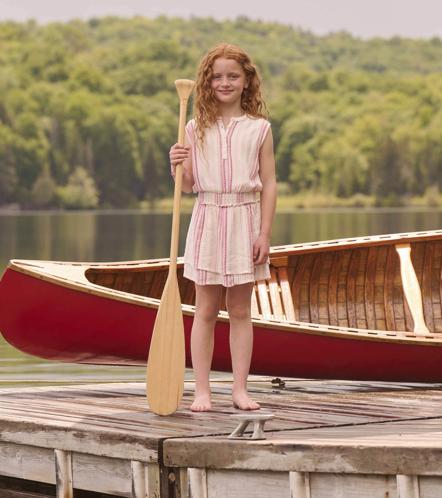 Light pink dress with ribbon stripes and smocked waist, worn by a girl standing on a dock holding a wooden paddle.