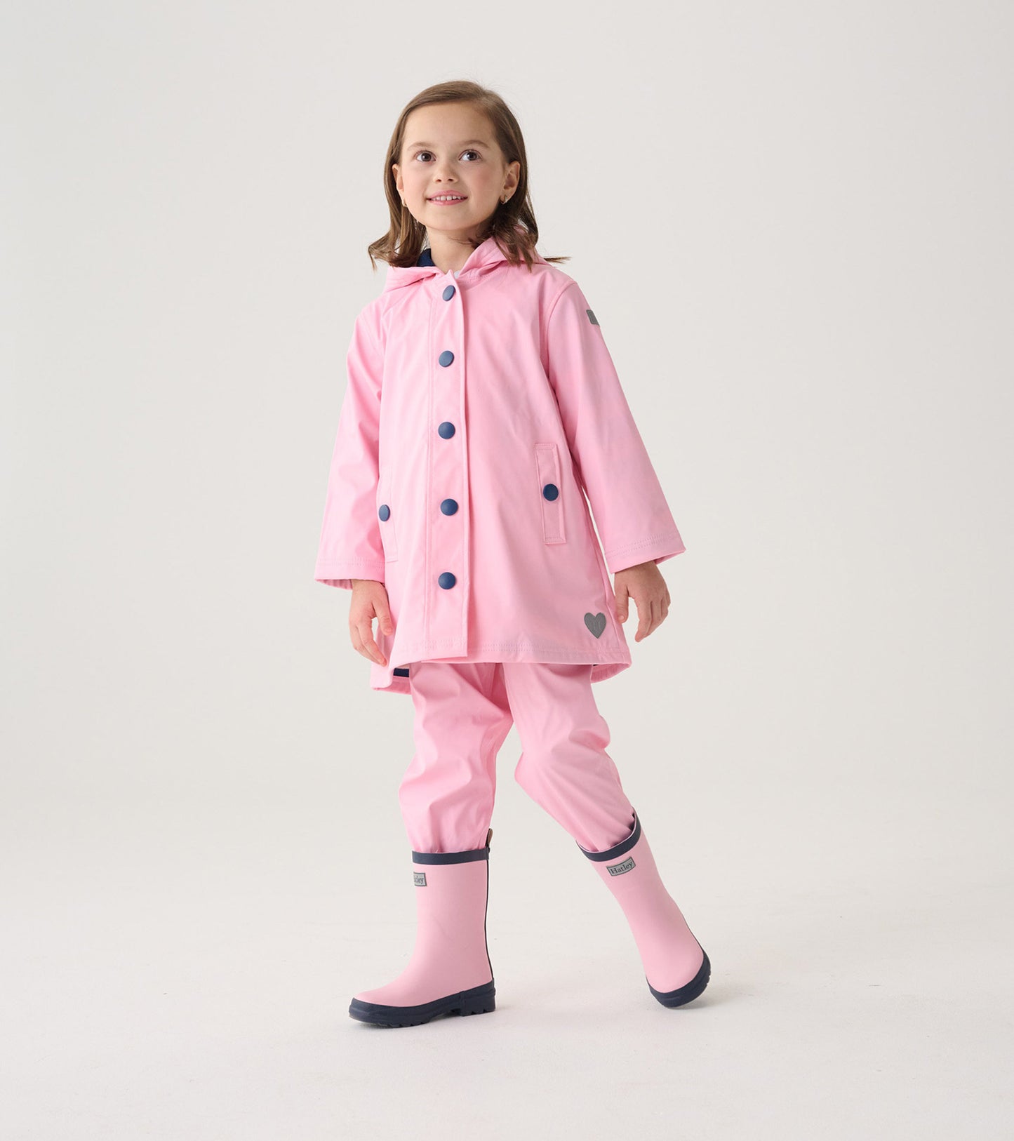 Pink button-up rain jacket with navy buttons, hood, and heart detail, worn by a girl with matching rain boots.