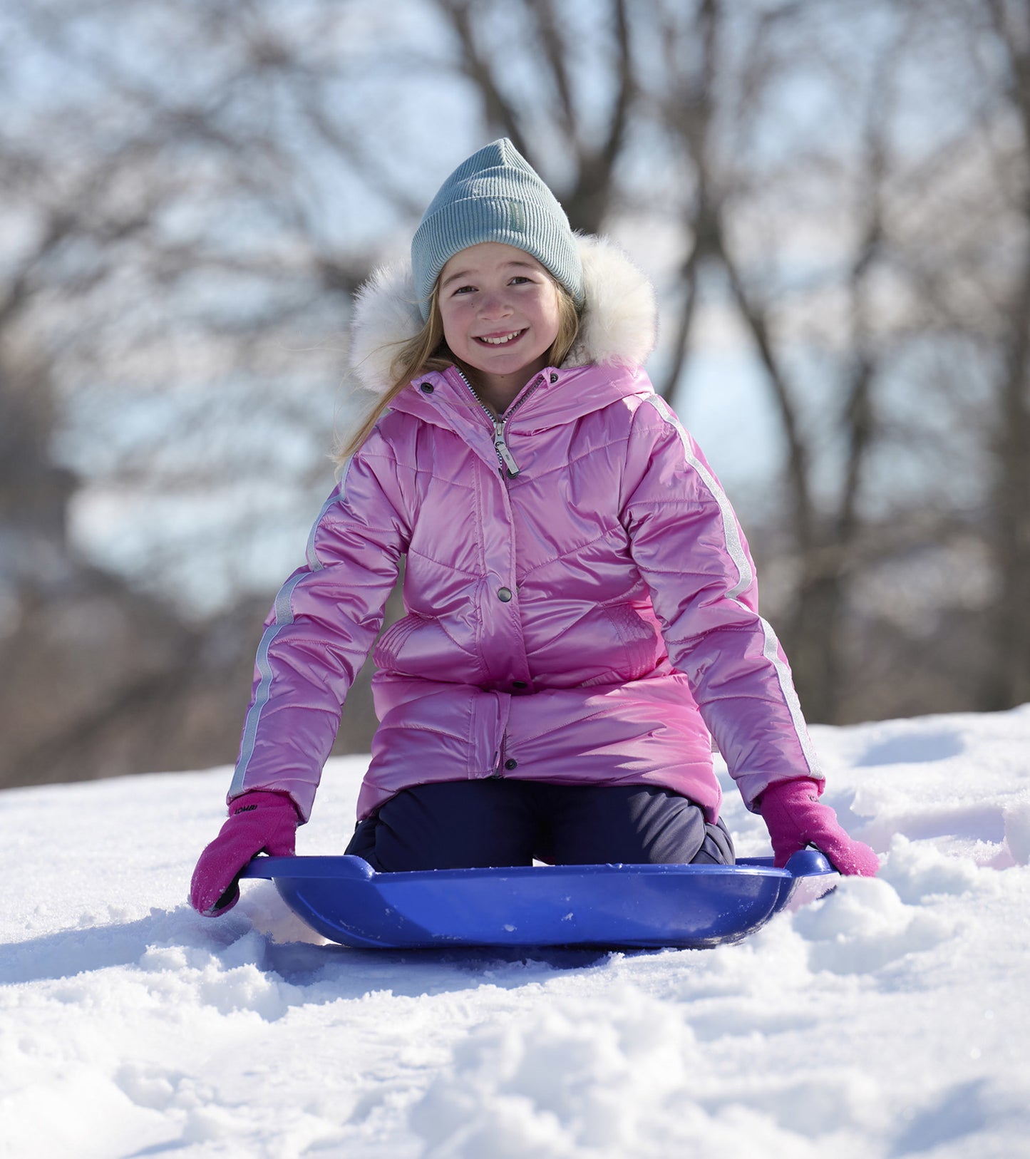 Pink puffer jacket with fur trim, worn by a girl sitting on a blue sled in the snow, smiling in a winter landscape.