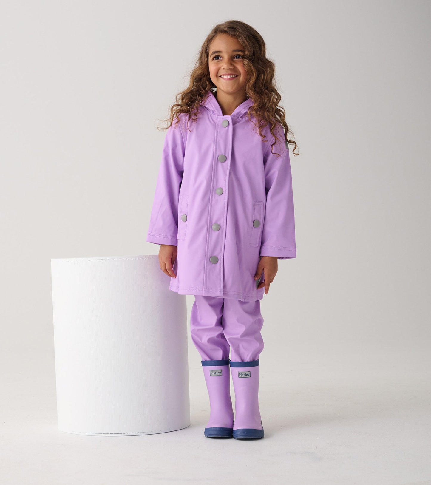Lilac button-up rain jacket with a hood and gray buttons, worn by a girl standing next to a white cylindrical object.