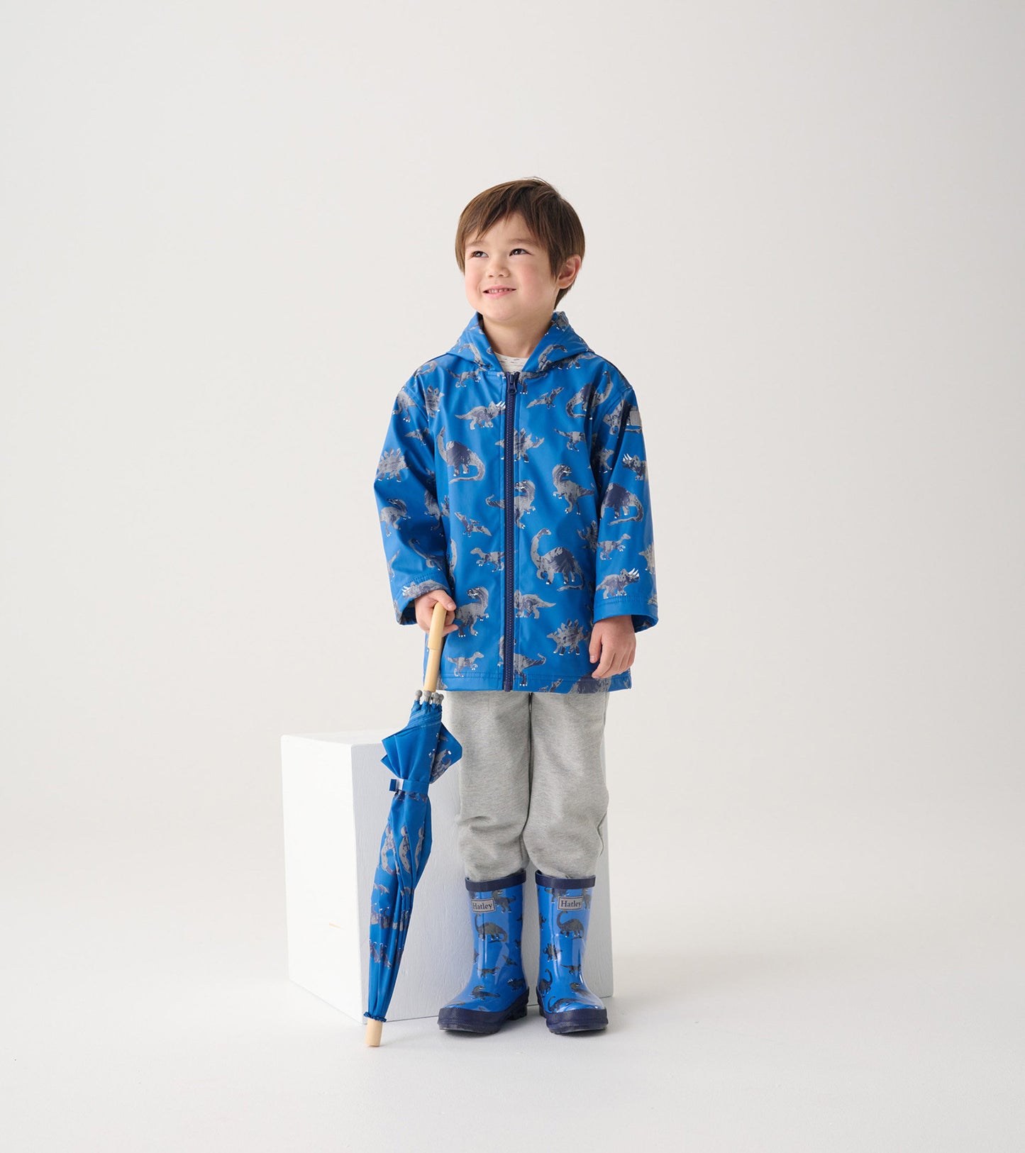 Blue zip-up rain jacket with dinosaur print, worn by a boy holding a matching umbrella, paired with gray pants and rain boot…