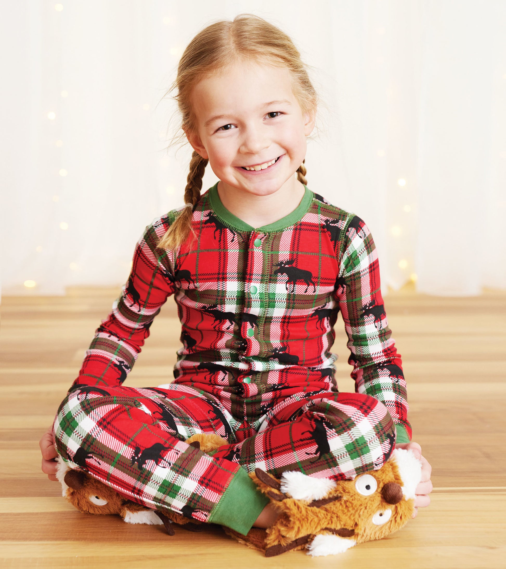Red plaid kids' onesie featuring moose patterns, with green trim, worn by a smiling girl sitting cross-legged.