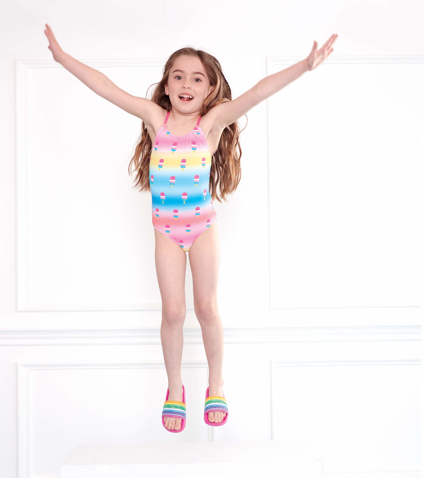Colorful slides with rainbow stripes, worn by a girl jumping in a swimsuit with ice cream print.