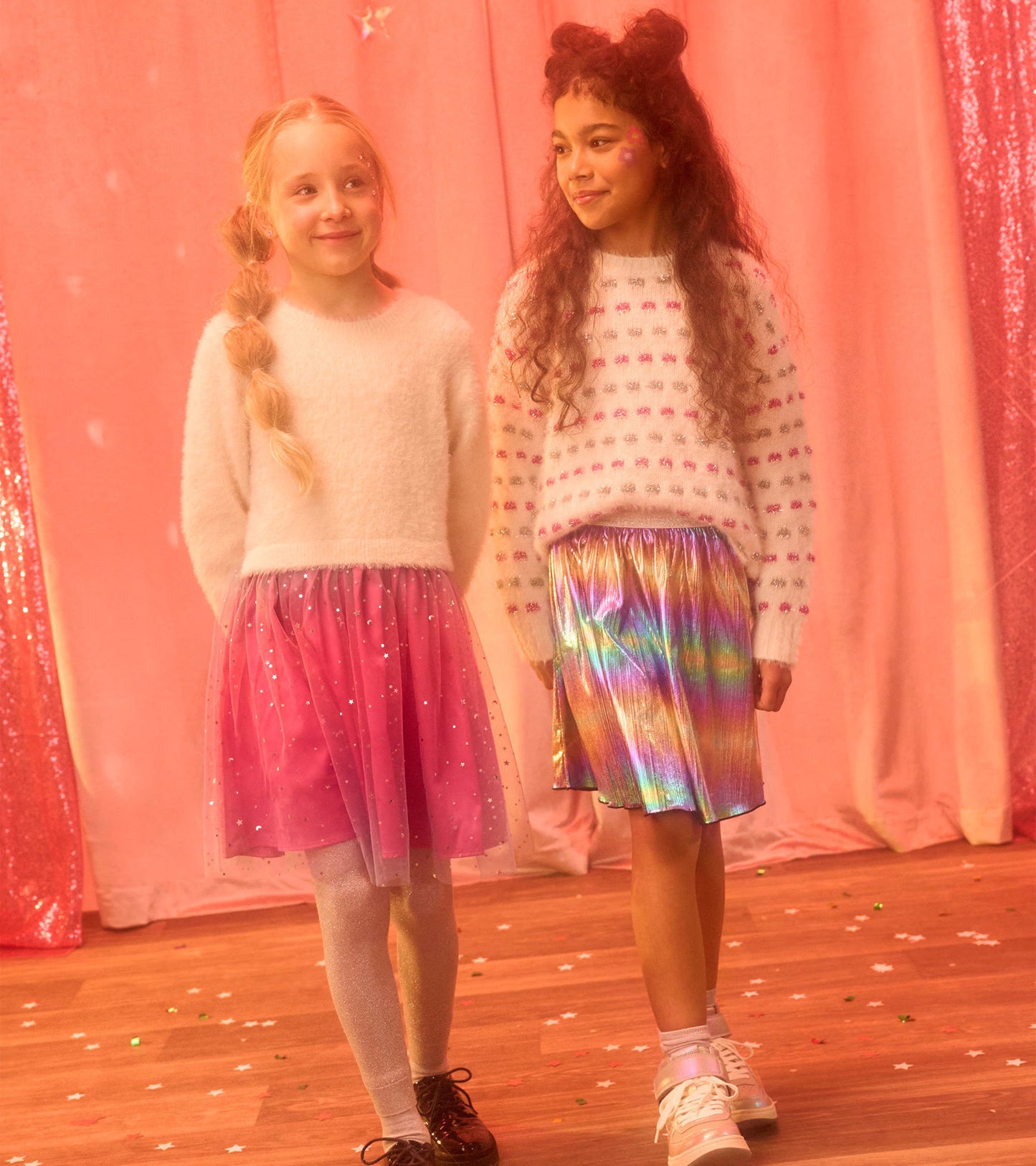 White sweater tunic with colorful dots, paired with a pink tulle skirt and a shiny rainbow skirt, worn by two girls.