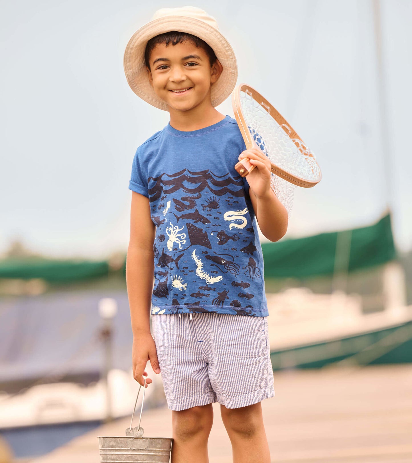 Blue graphic tee featuring ocean-themed designs, worn by a child holding a net, paired with striped shorts.