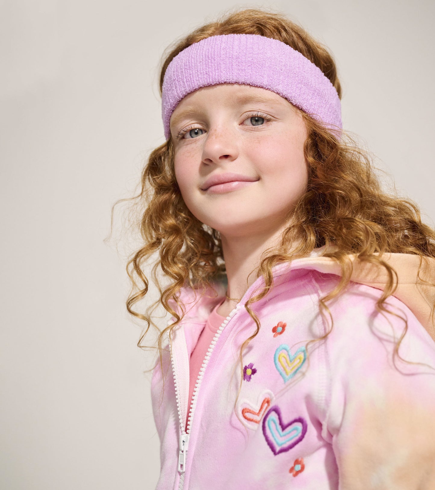 Pink tie-dye hoodie with heart and flower embroidery, featuring a zip front and hood, worn by a girl with curly hair.