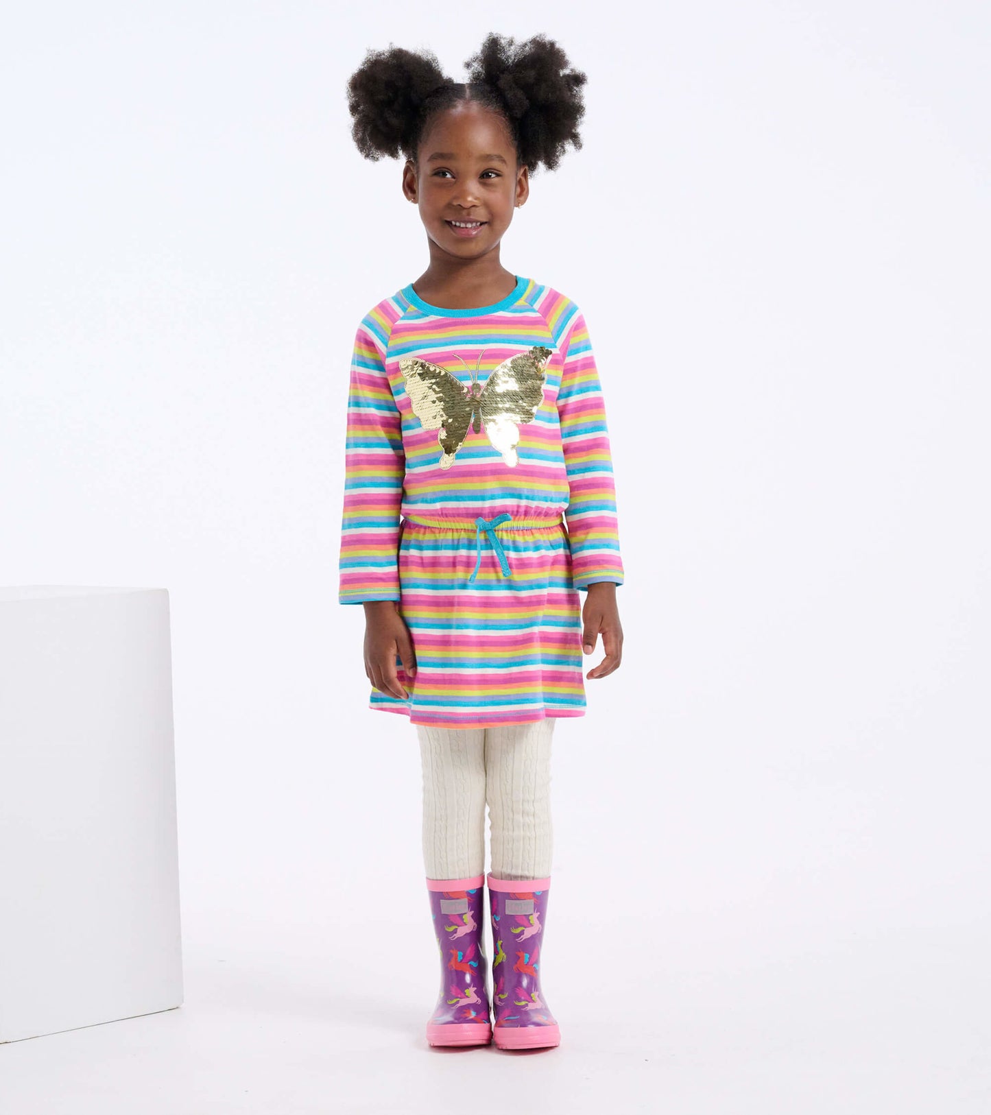 Colorful striped drop waist dress with a butterfly design, paired with cream leggings and pink rain boots.
