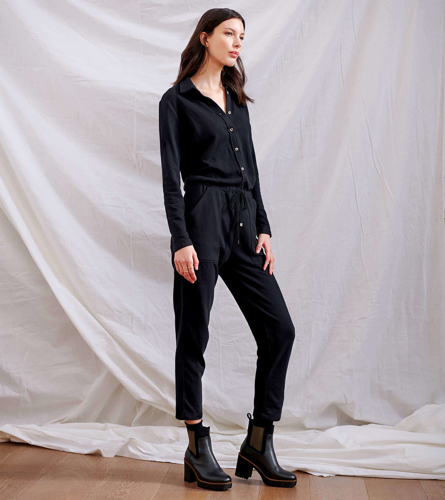 Black long-sleeve jumpsuit with a button-up front, drawstring waist, and tapered legs, styled with ankle boots.