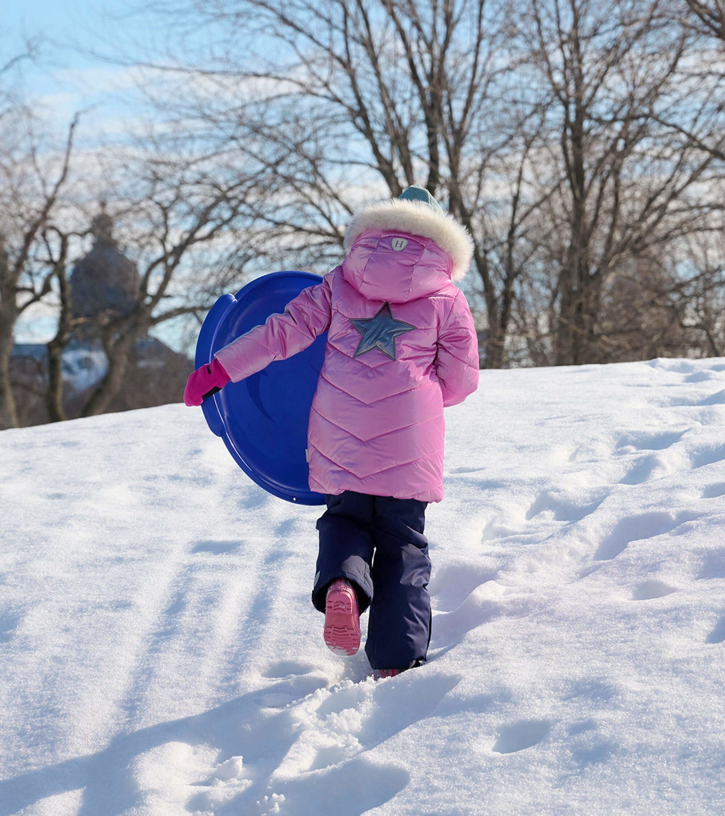 Pink puffer jacket with a star design, worn by a child carrying a blue sled while walking in the snow.