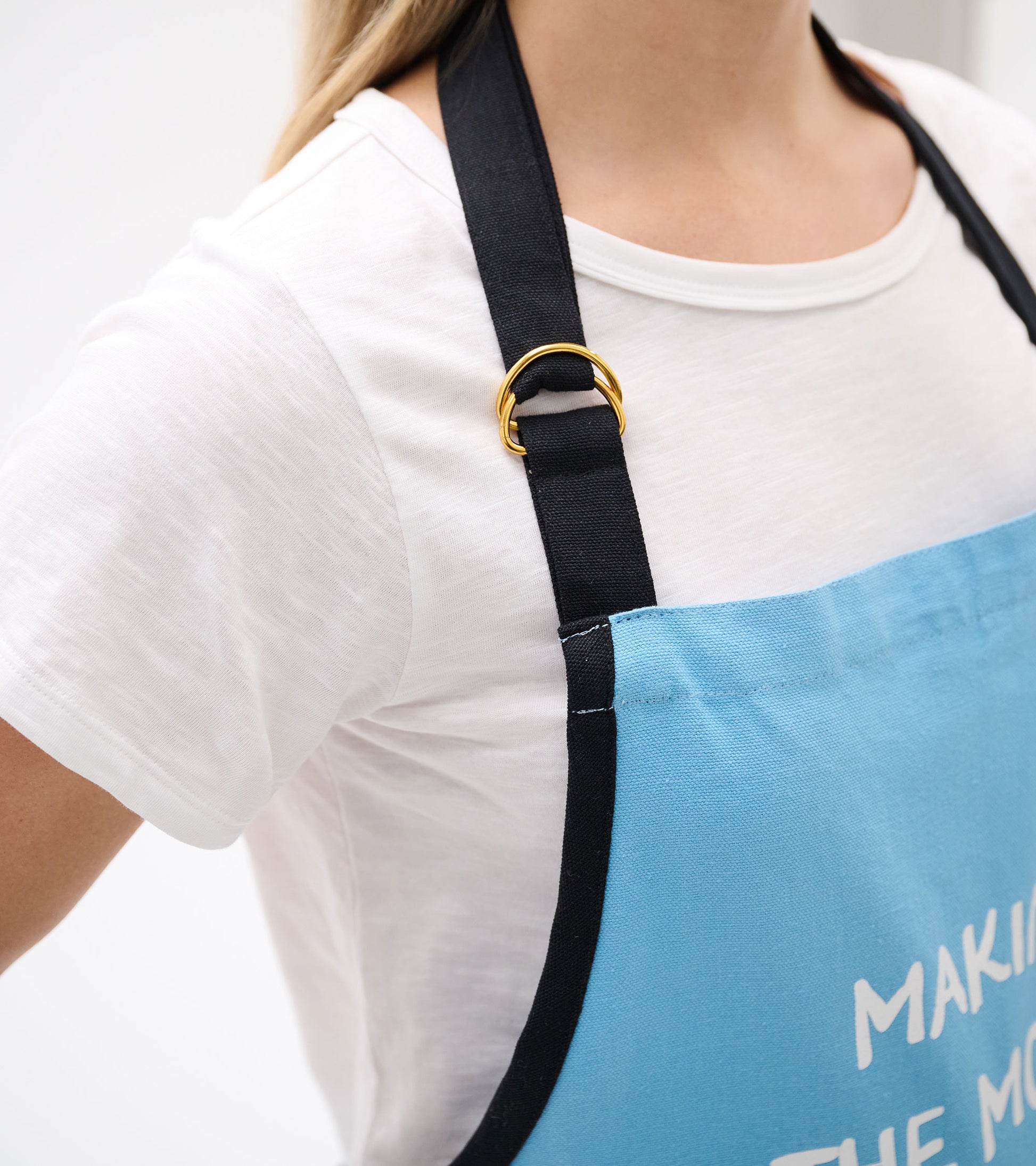 Light blue apron with black straps and a gold ring detail, worn over a white shirt, showcasing a casual cooking style.