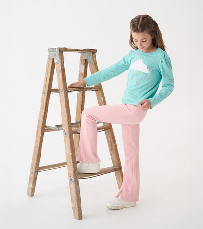 Light blue long-sleeve tee with a fluffy cloud graphic, paired with pink flared pants, worn by a girl on a ladder.