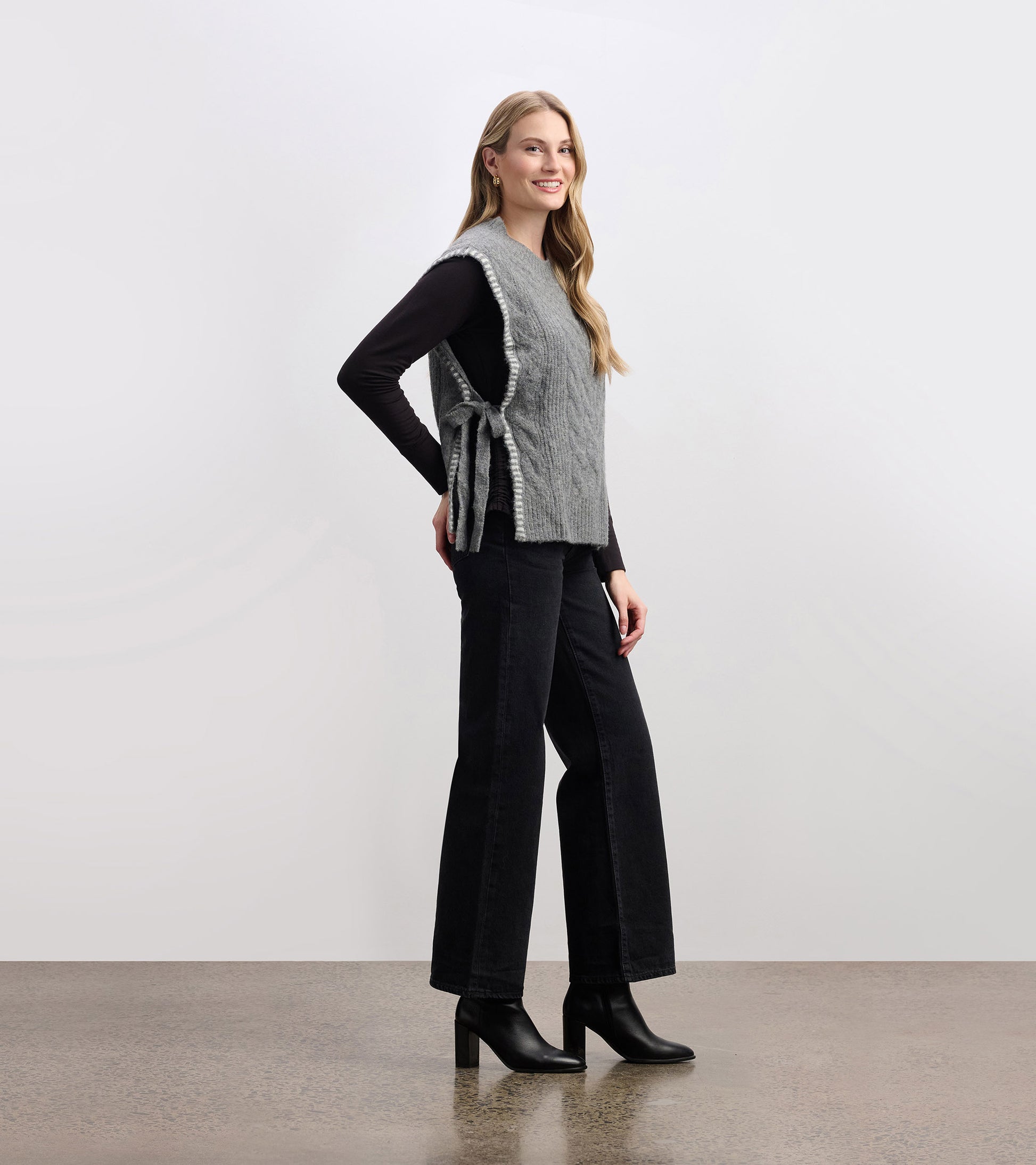 Charcoal melange side-tie sweater vest with a textured knit, styled over a black long-sleeve top and paired with wide-leg pa…