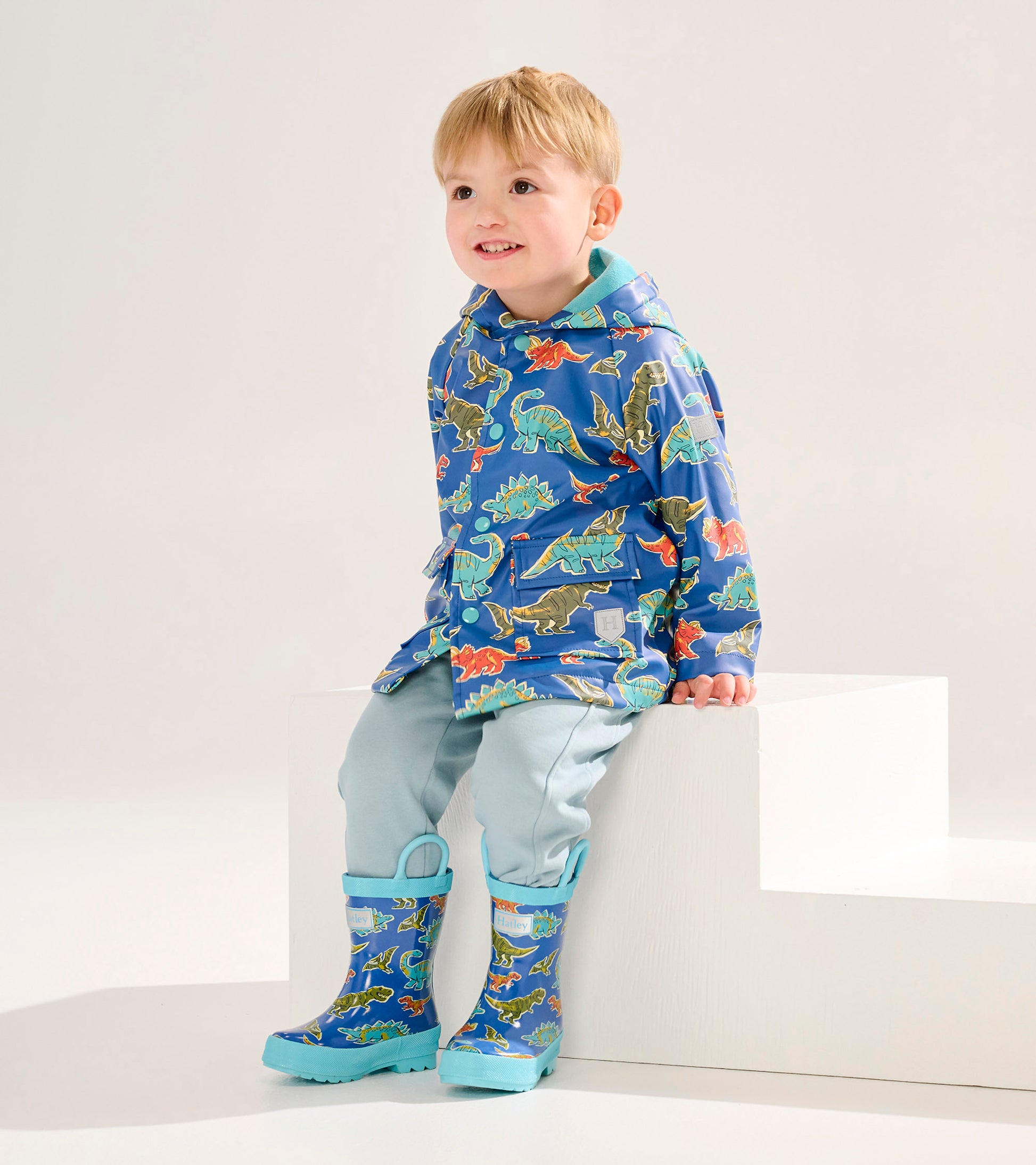 Blue dinosaur-patterned rain jacket on a smiling toddler, paired with light blue pants and matching rain boots.