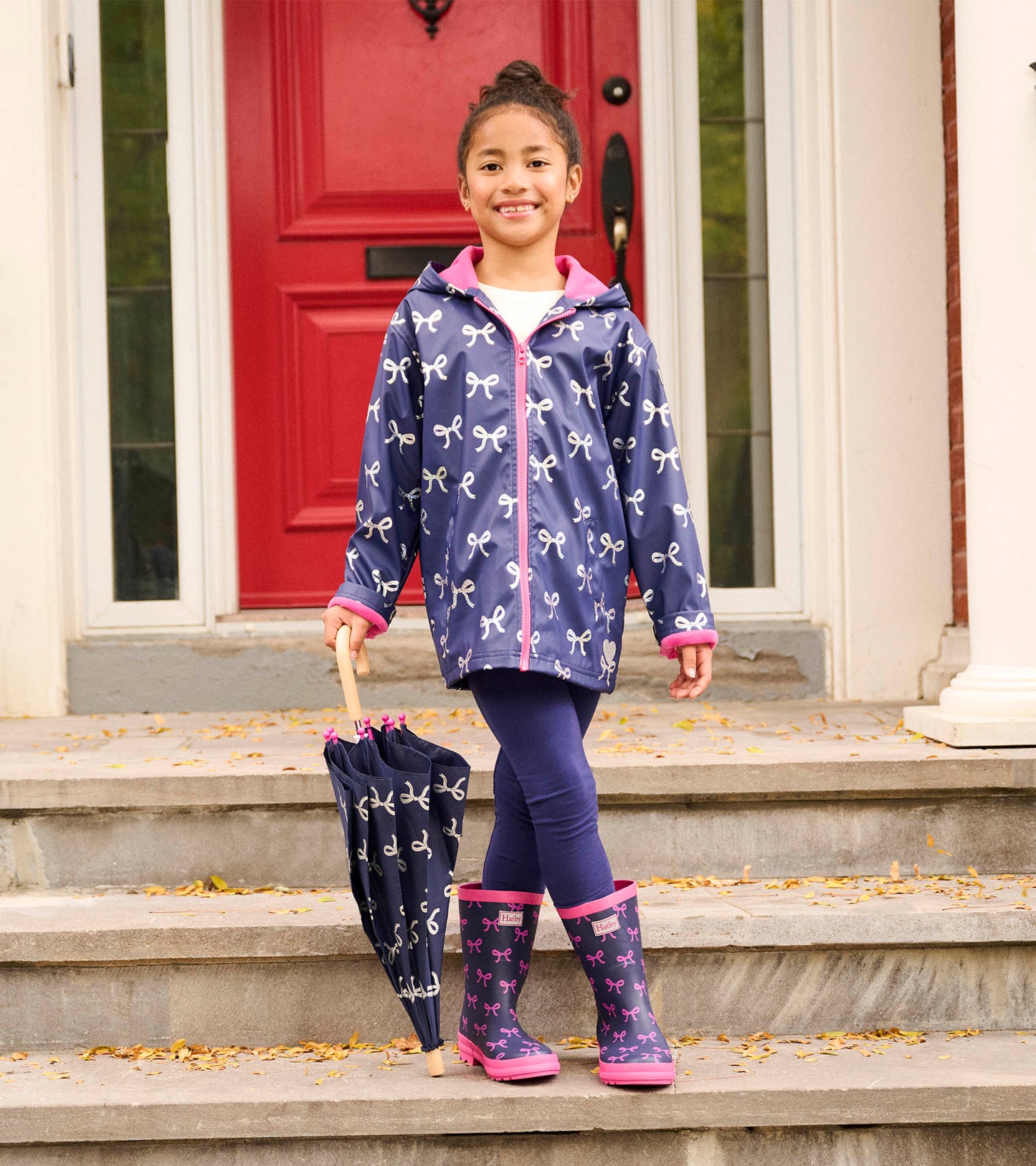 Navy rain jacket with white bows, pink trim, and hood, worn by a girl holding a matching umbrella and wearing rain boots.