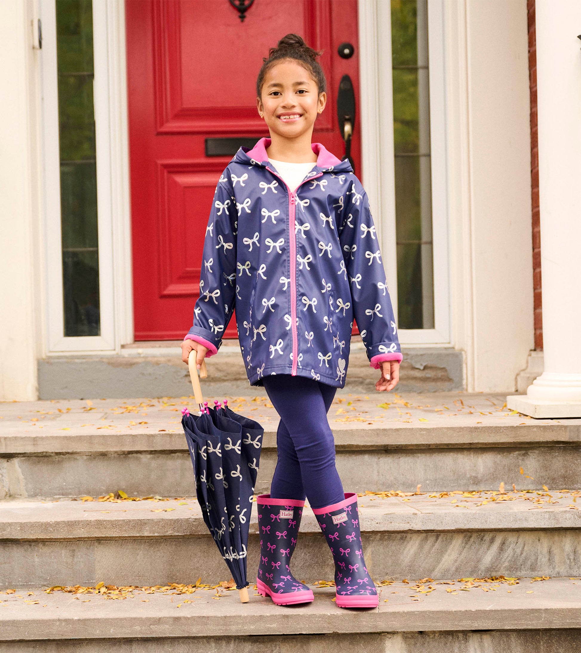 Navy rain jacket with white bows, pink trim, and hood, worn by a girl holding a matching umbrella and wearing rain boots.