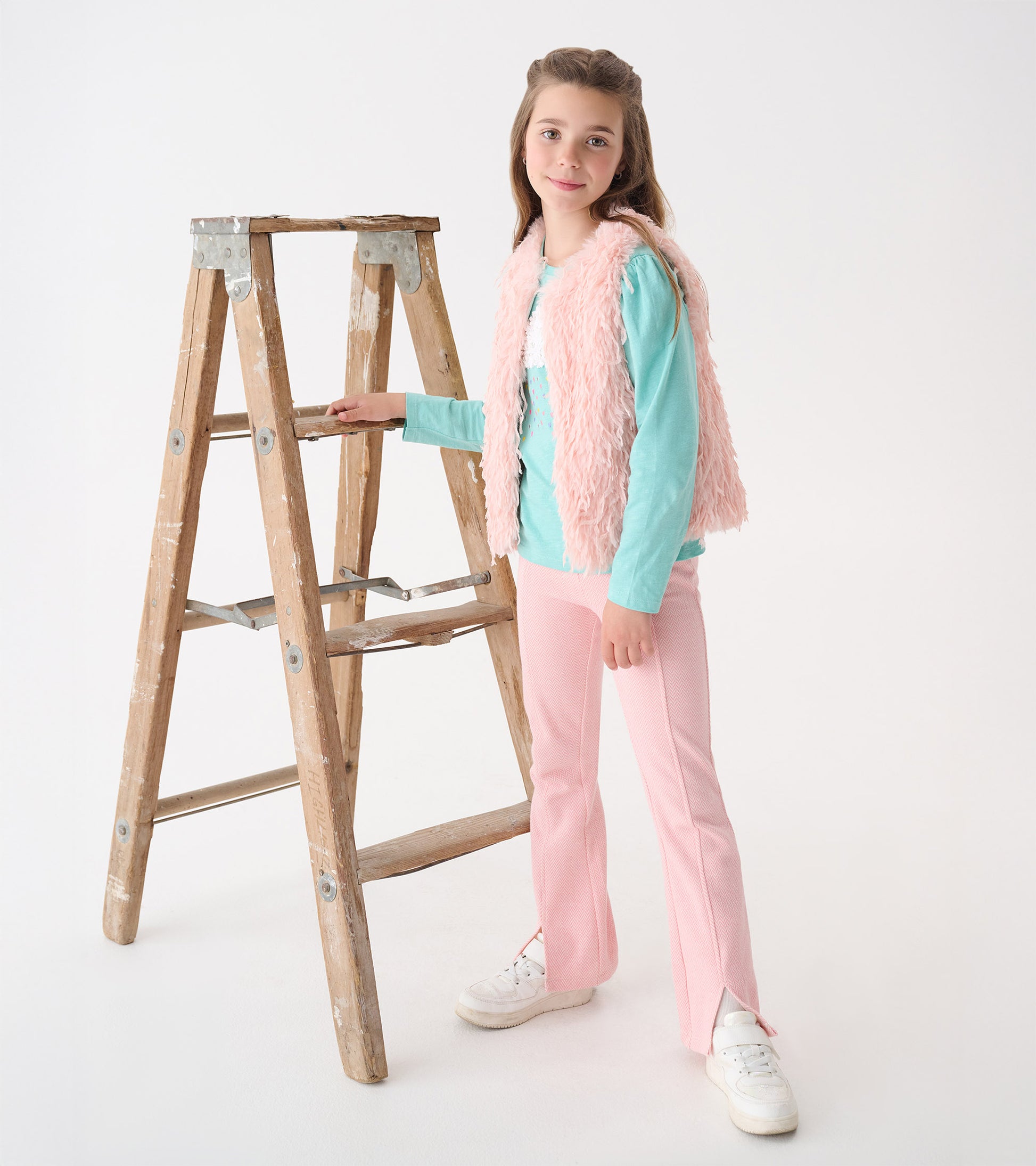 Pale pink faux fur vest worn over a light blue top, paired with pink pants and white sneakers, standing by a wooden ladder.