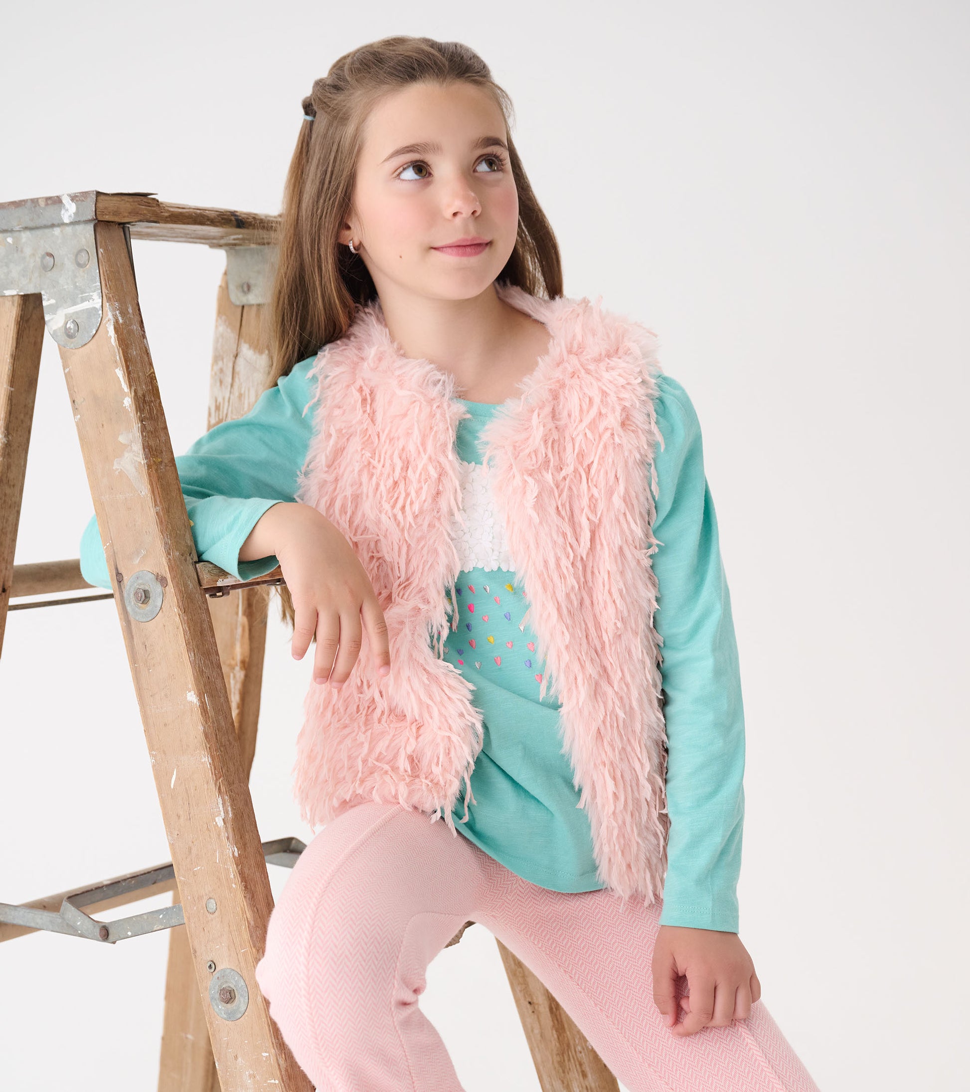 Pale pink faux fur vest worn over a teal top, paired with light pink pants, on a girl sitting beside a wooden ladder.