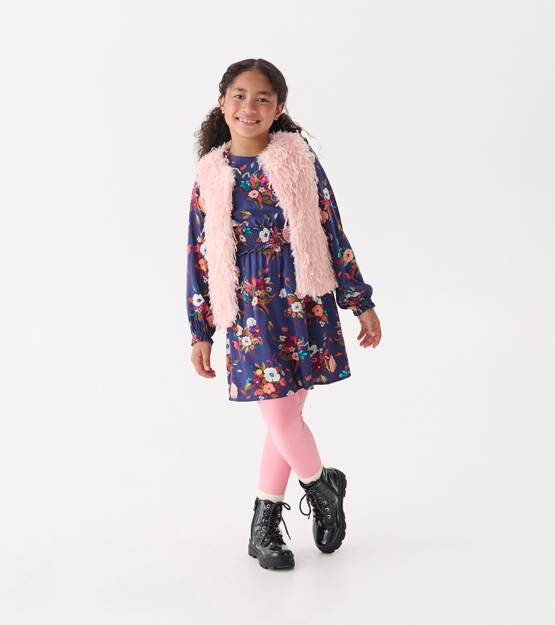 Pale pink faux fur vest worn over a floral dress, paired with pink leggings and black boots, on a smiling girl.