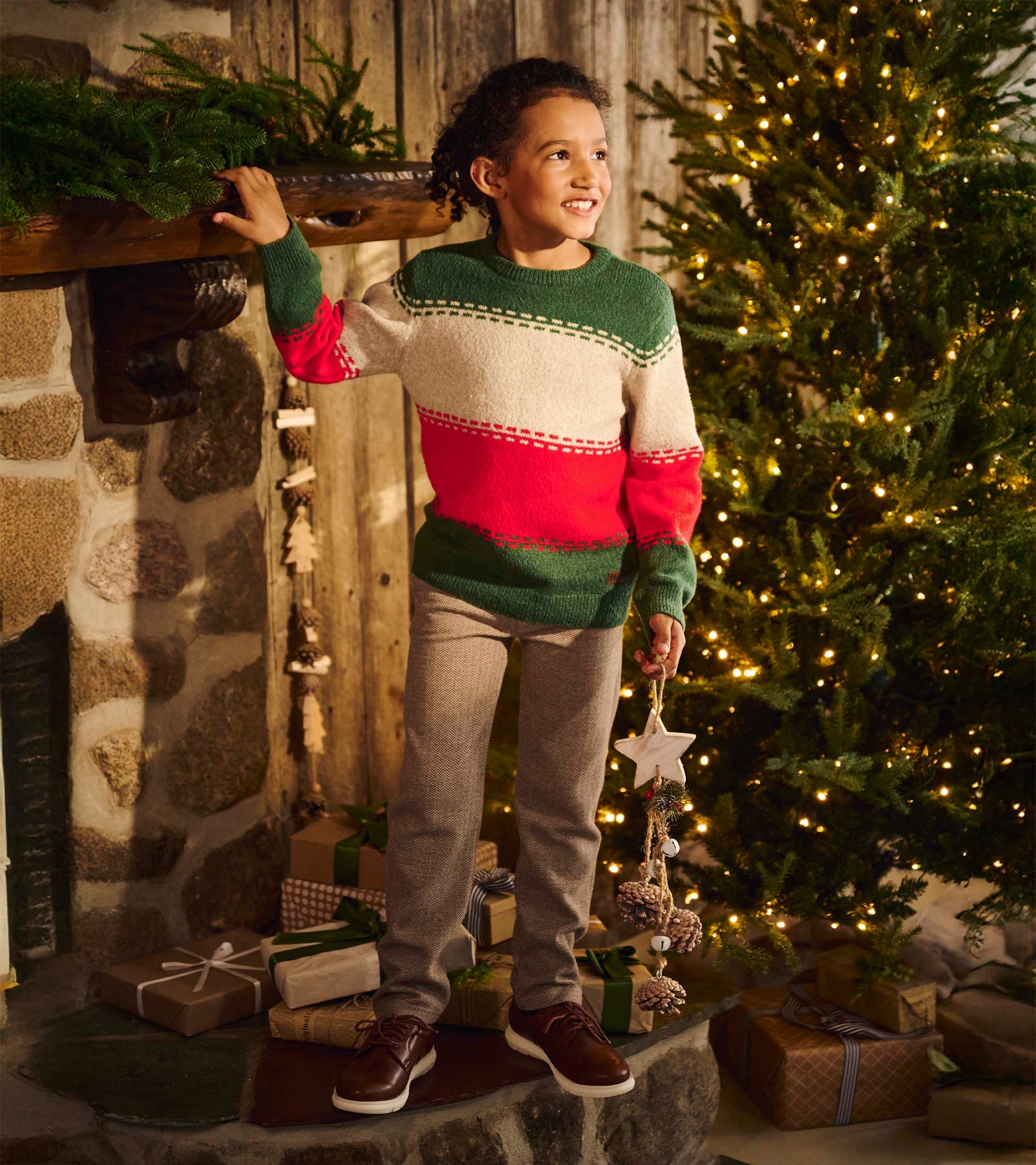 Boy wearing a green, cream, and red striped holiday sweater with gray pants by a lit Christmas tree