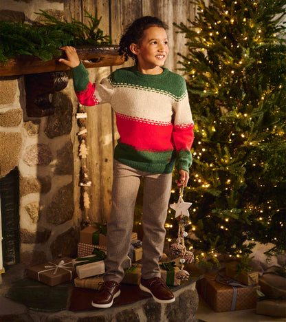 Boy wearing a green, cream, and red striped holiday sweater with gray pants by a lit Christmas tree