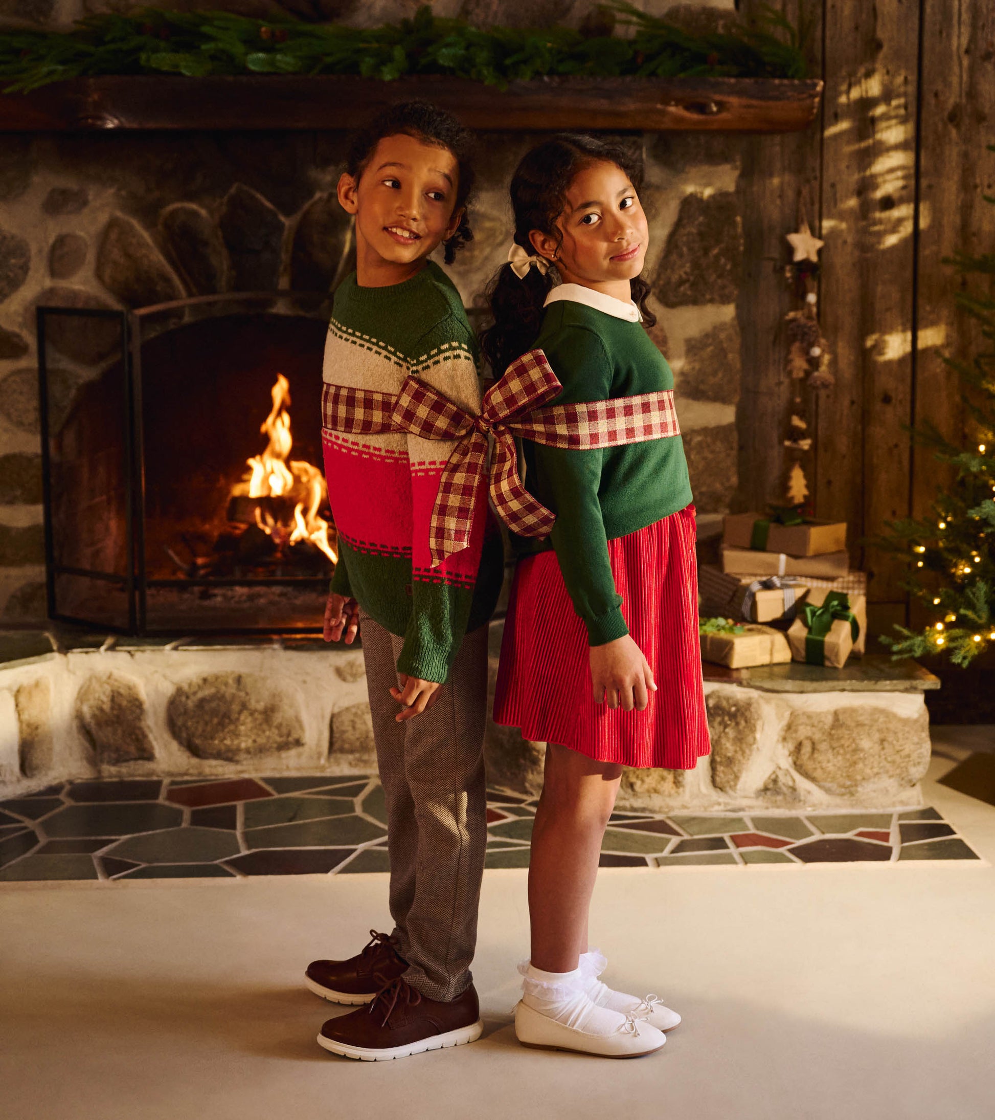 Two children wearing holiday sweaters tied together with a plaid ribbon, standing by a fireplace