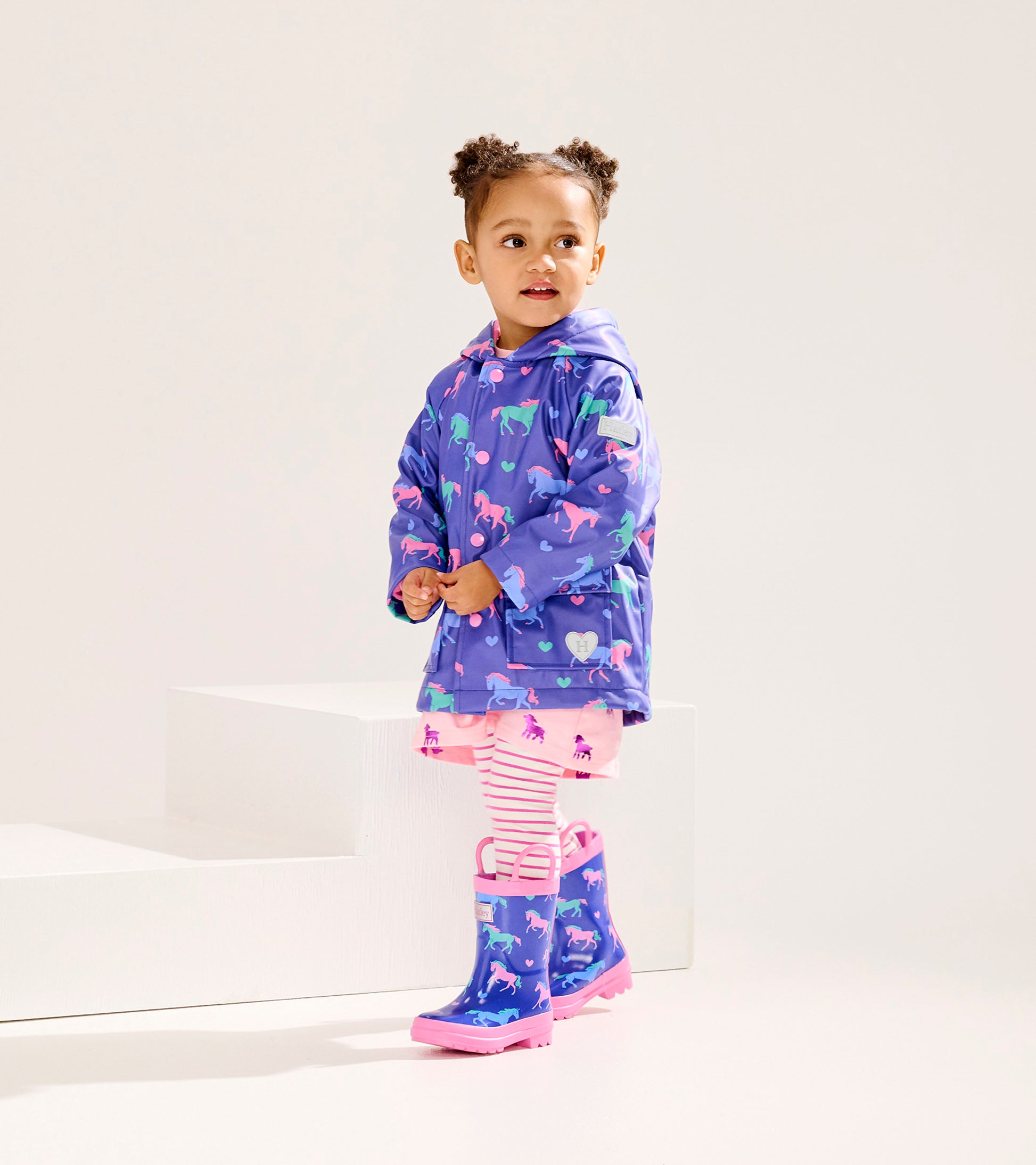 Purple rain jacket with horse and heart patterns, worn by a girl with pink rain boots and striped leggings.