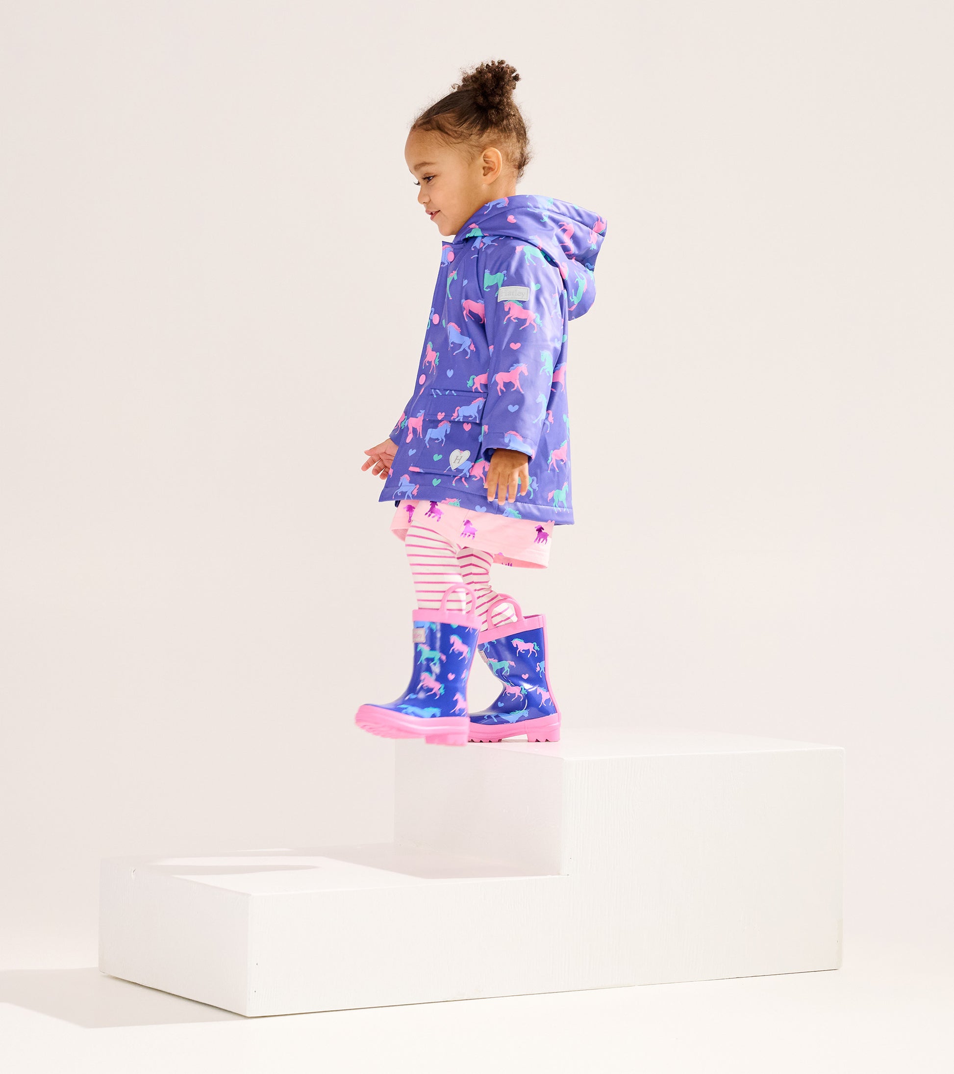 Purple rain jacket with horse and heart patterns, worn by a girl with striped leggings and matching rain boots.