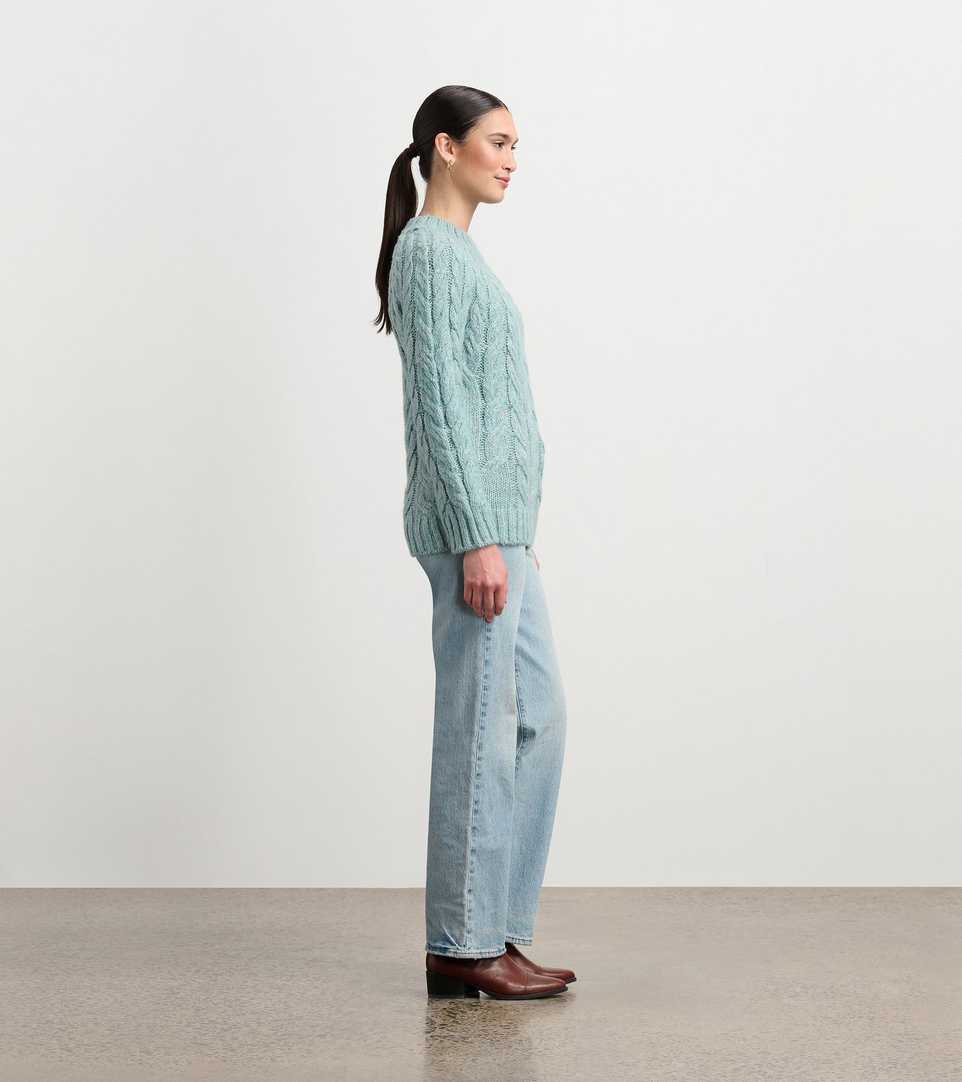 Light turquoise cable knit sweater with a relaxed fit, worn with light blue jeans and brown boots, viewed from the side.
