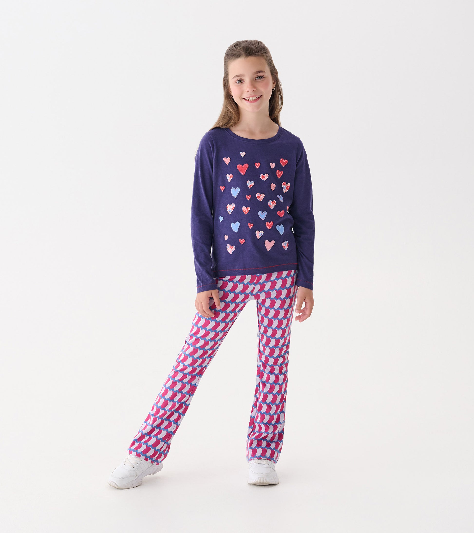Long sleeve purple tee with colorful heart graphics, paired with patterned pink and purple pants, worn by a smiling girl.