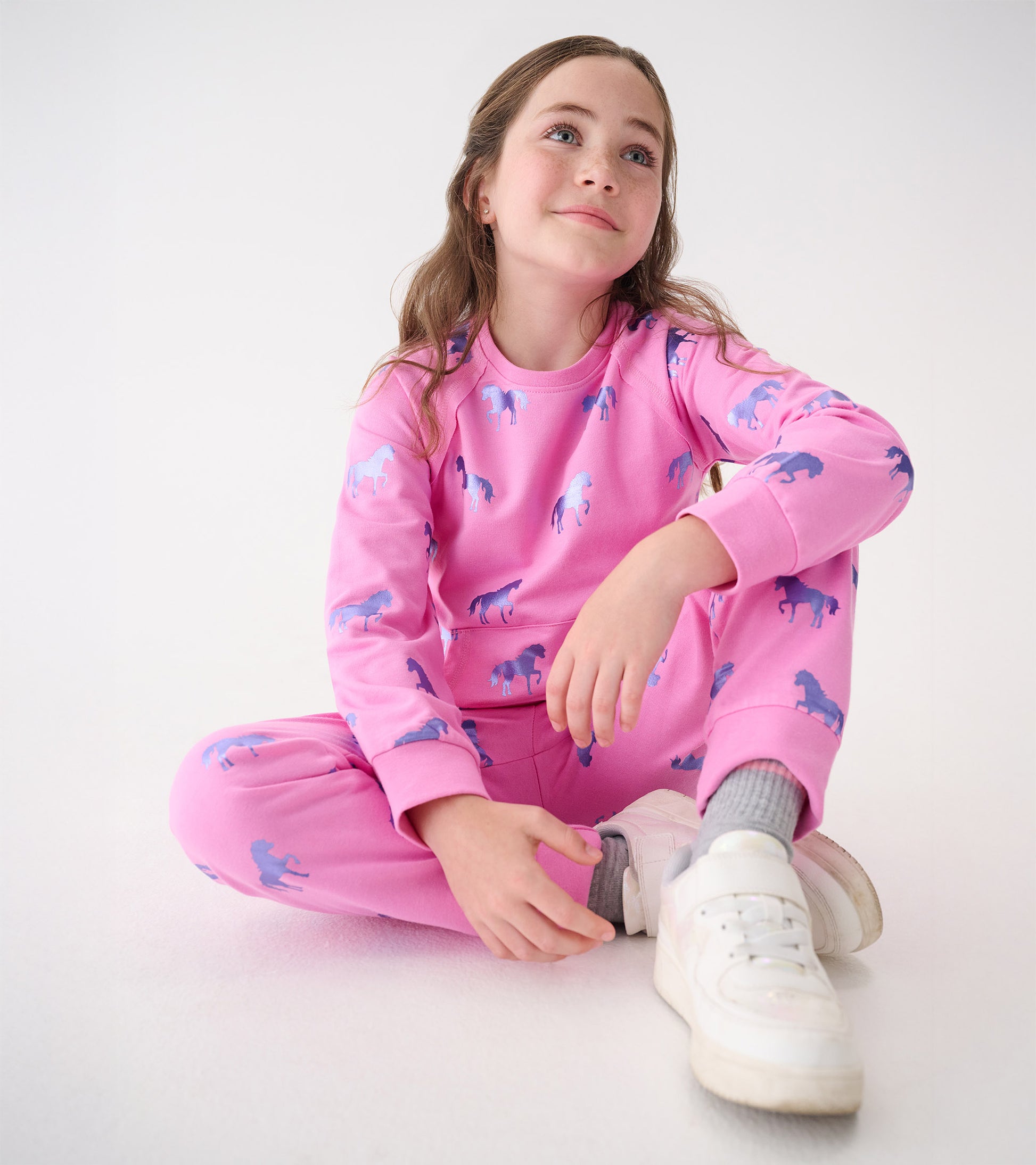 Pink sweater featuring a horse pattern, worn by a girl sitting cross-legged, paired with white sneakers.