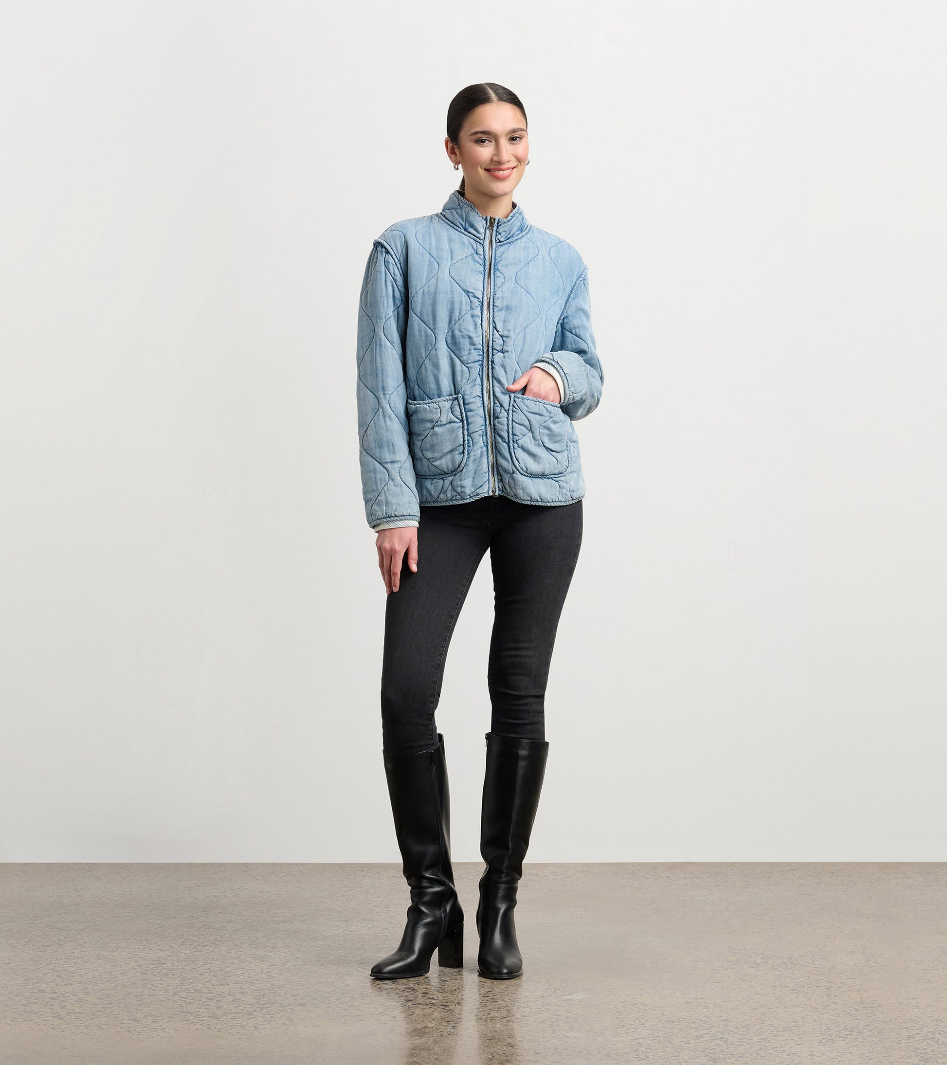 Quilted jacket in washed blue Tencel, featuring a high collar and front pockets, styled with black jeans and boots.