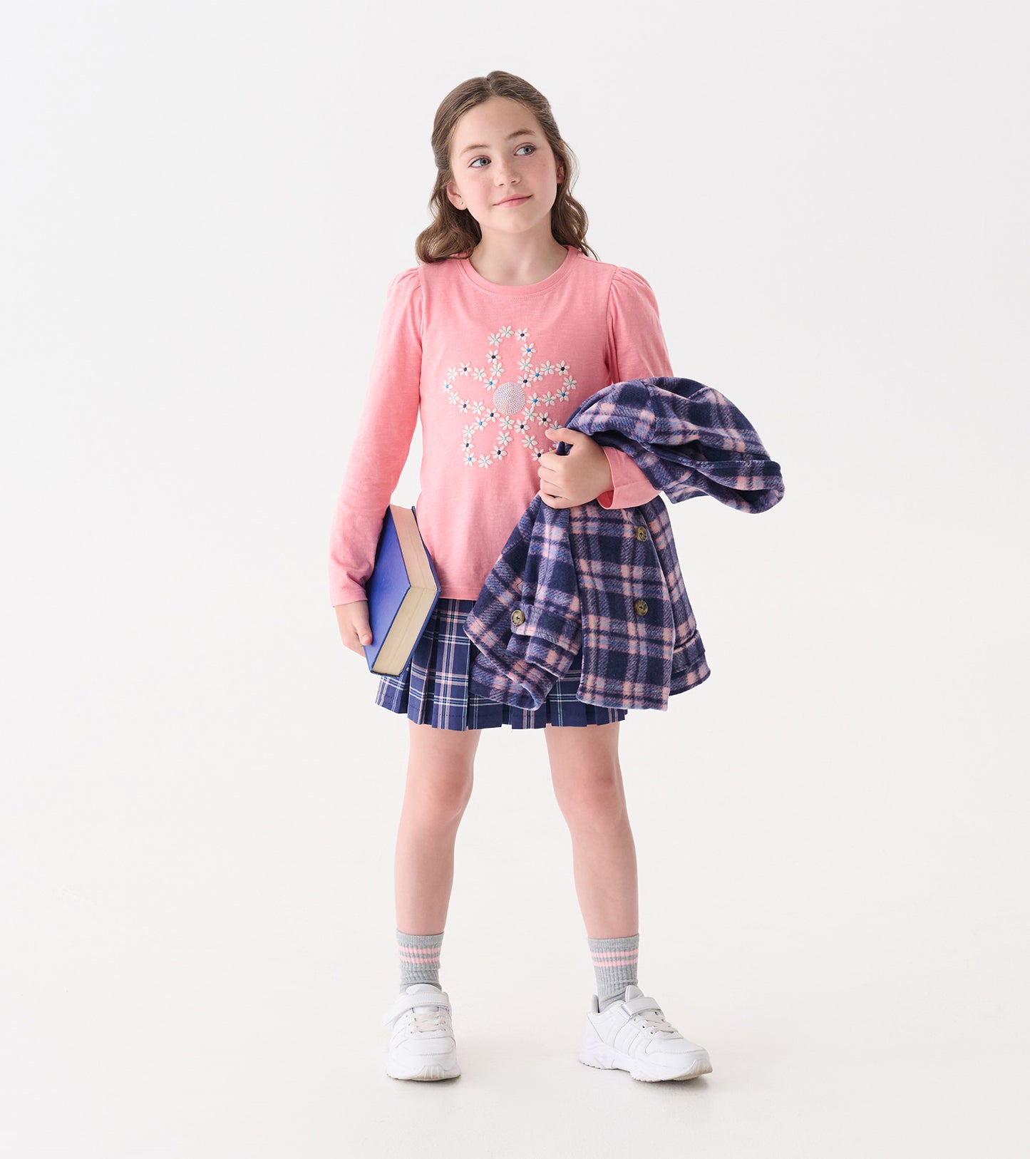 Pink long-sleeve tee with embroidered flower design, paired with a plaid skirt and holding a book and jacket.