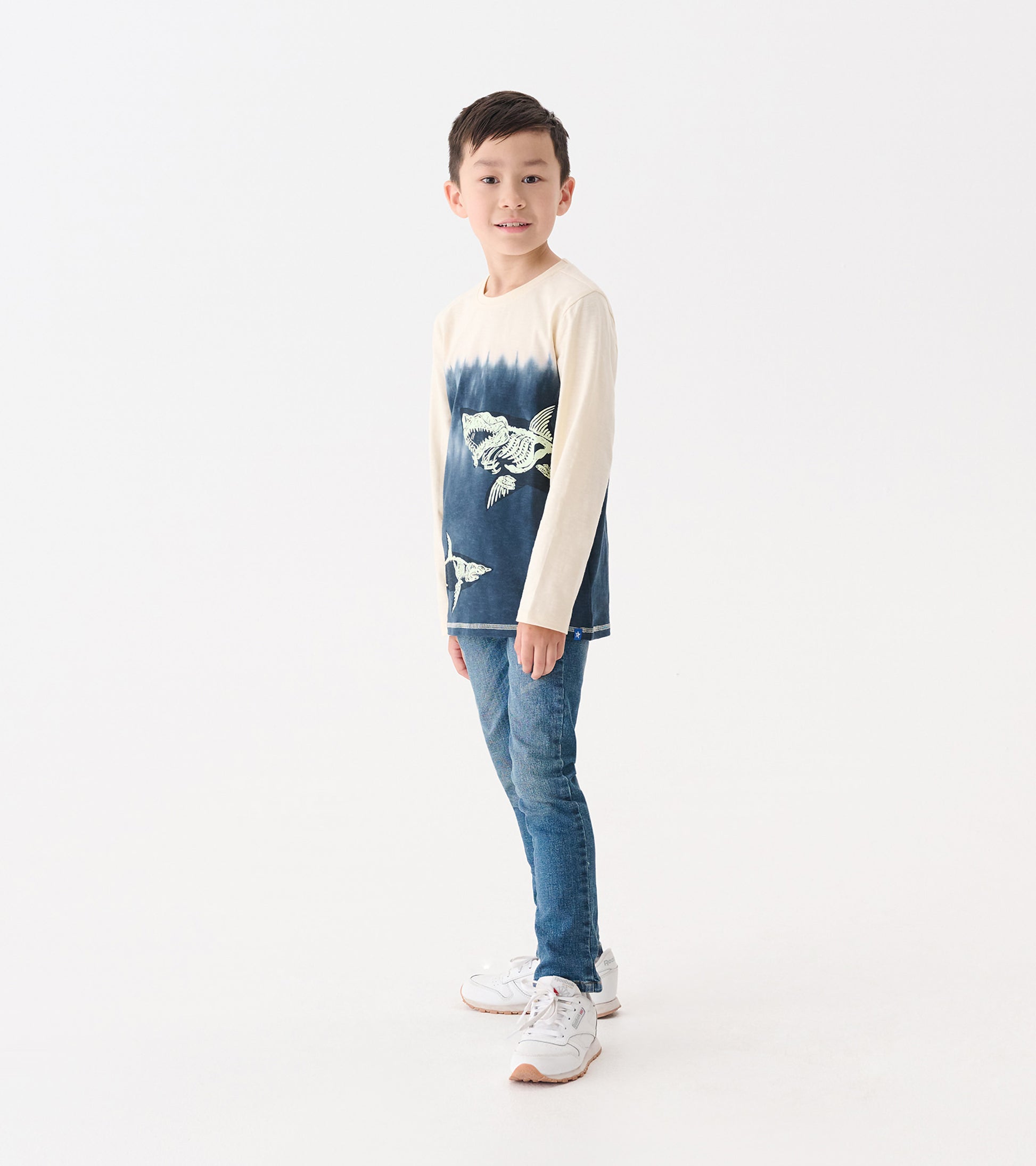 Long sleeve graphic tee featuring a shark design in blue and cream, worn by a boy with jeans and sneakers.