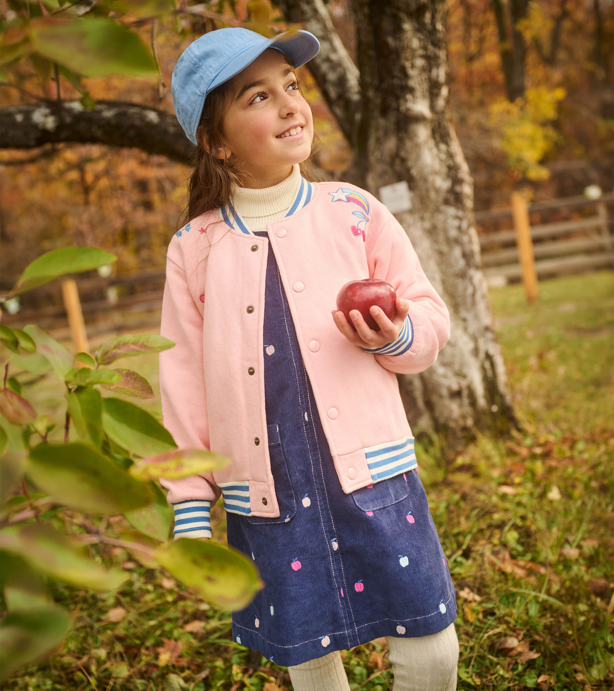 Girl wearing a navy pinafore dress with scattered apple embroidery, layered under a pink jacket outdoors