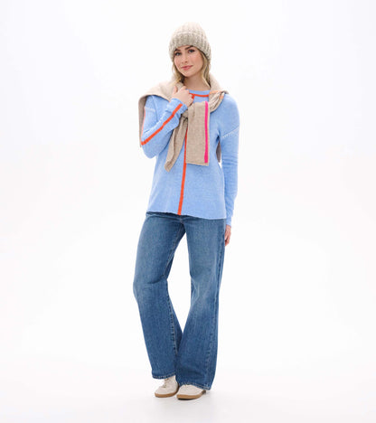 Front view of Maeve sweater in azure blue with orange trim and seam details, styled with jeans and a scarf