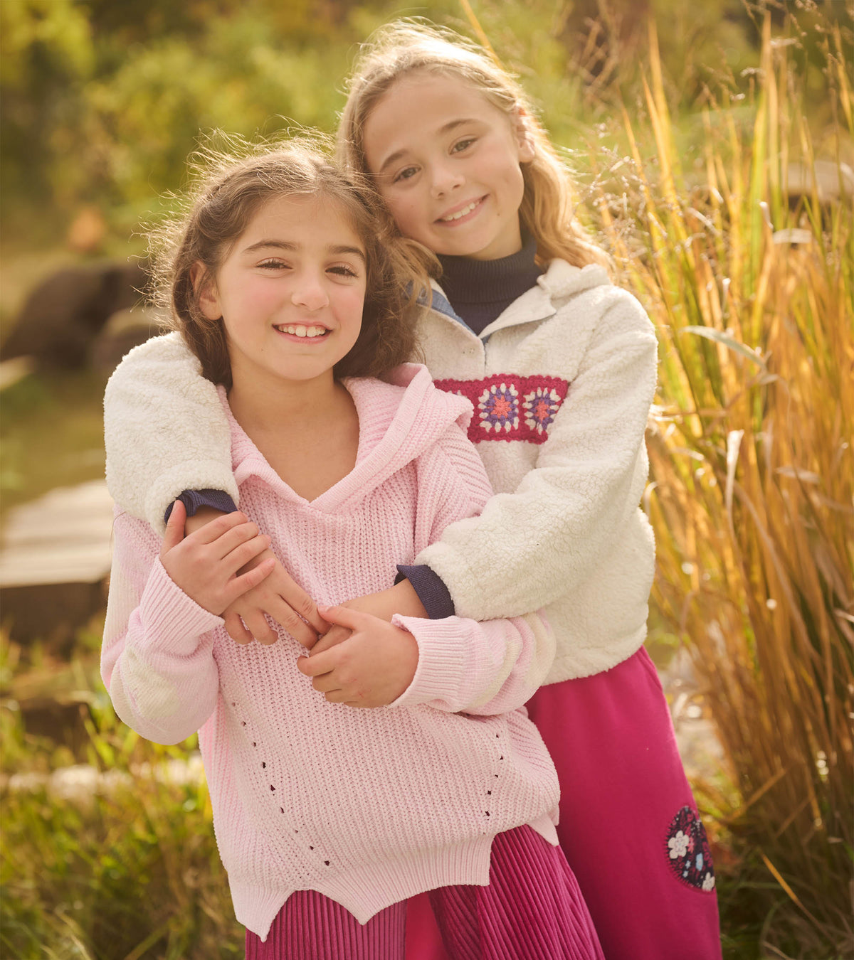 Girl wearing cream fuzzy pullover with crochet chest band, standing outdoors behind another child