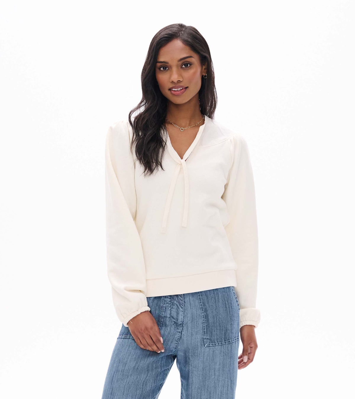 Front view of cream long sleeve top with tie neckline and banded hem, worn with blue pants