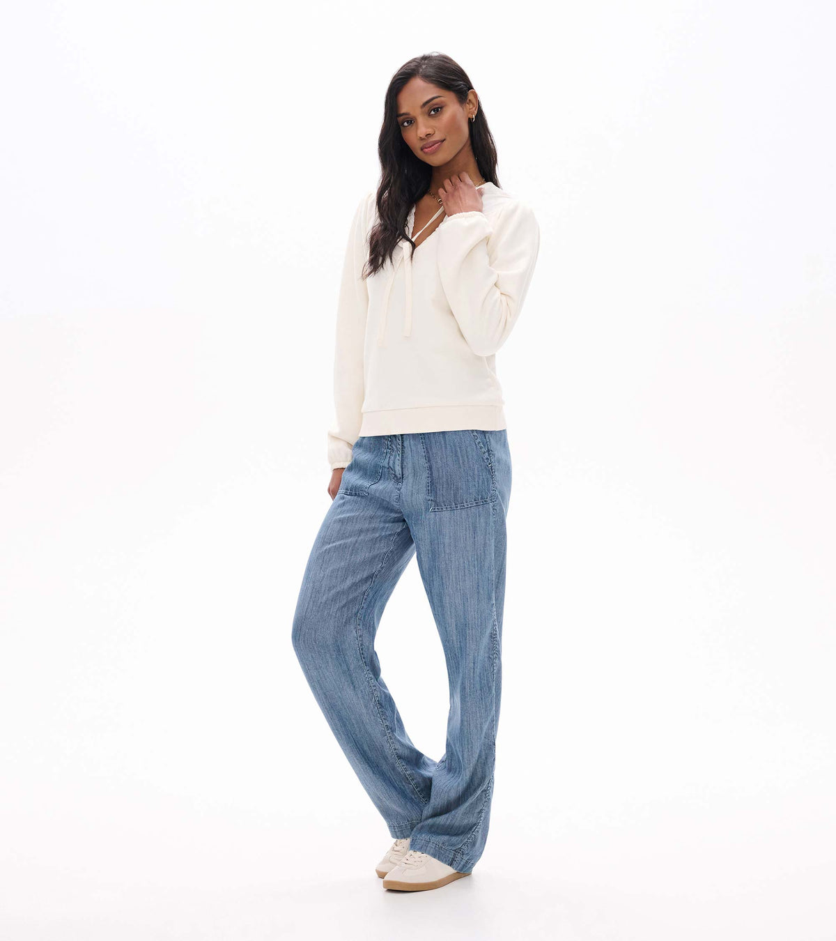 Three-quarter view of cream tie-neck long sleeve top with banded hem, worn with wide leg blue pants