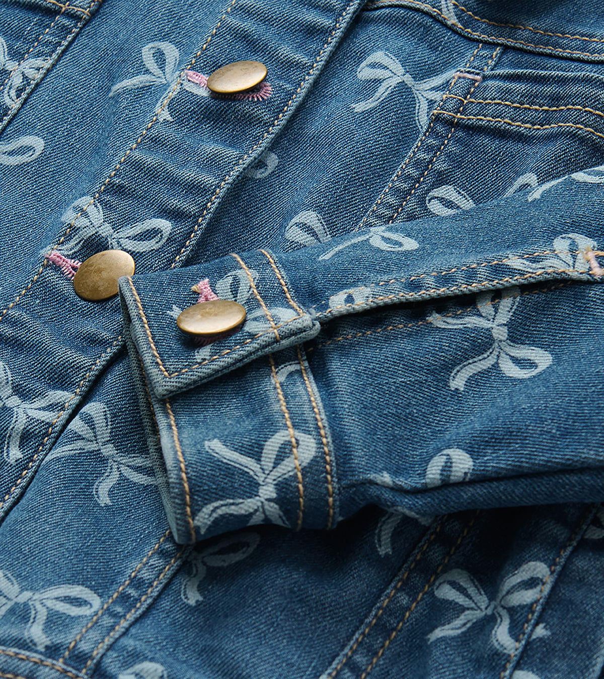 Close-up of blue bow-print denim jacket showing brass-tone buttons, cuff tab, and chest pocket detail
