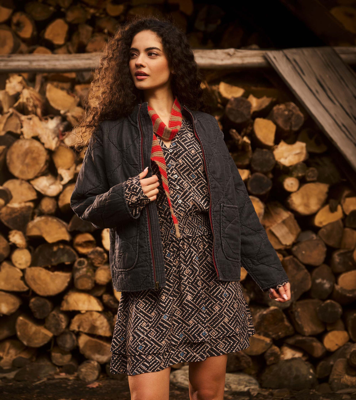 Model wearing black quilted jacket with burgundy zip, shown outdoors against stacked logs