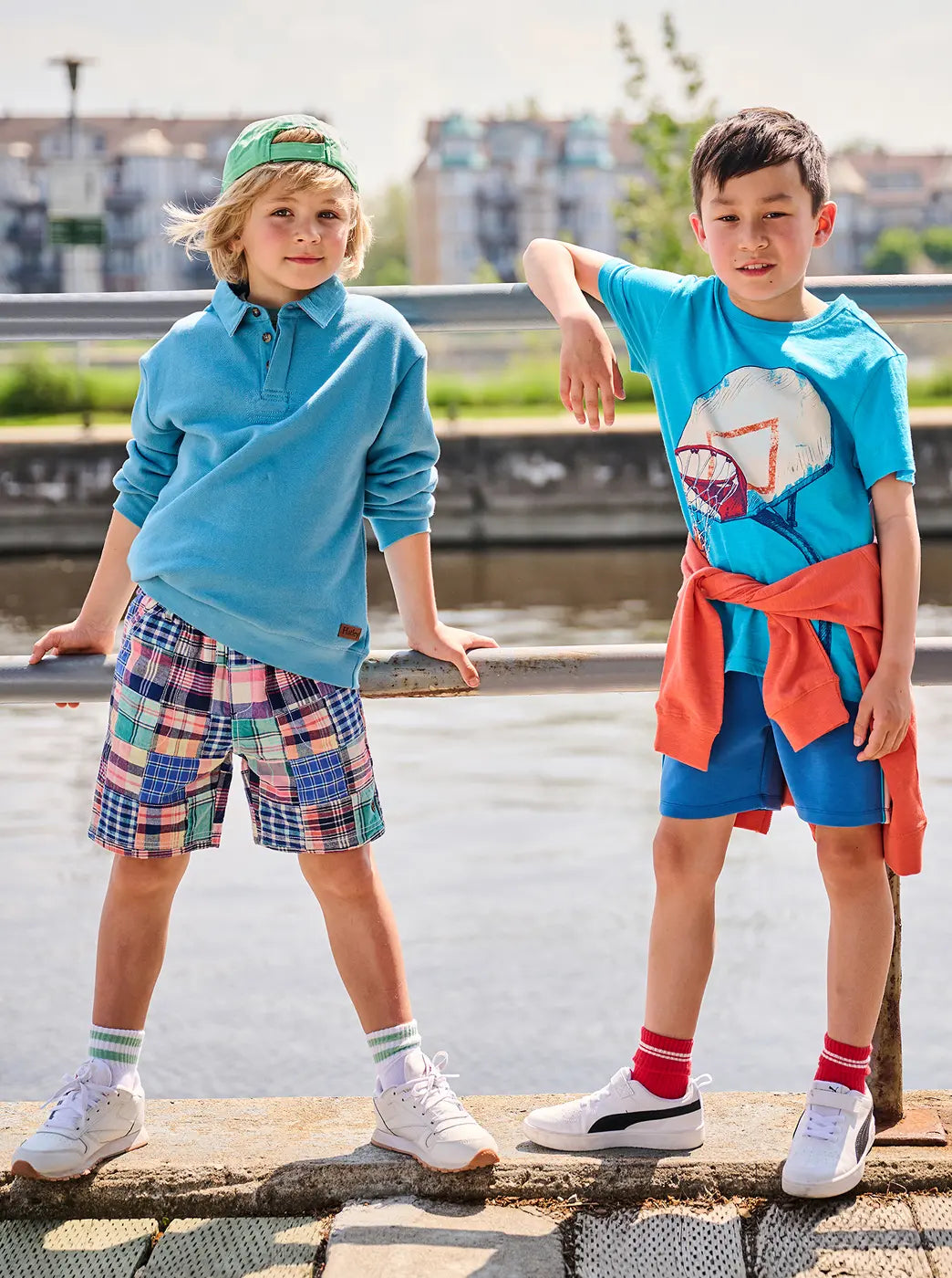 Shop boys' clothing collection. Showing two boys modeling colorful casual outfits by a riverside.