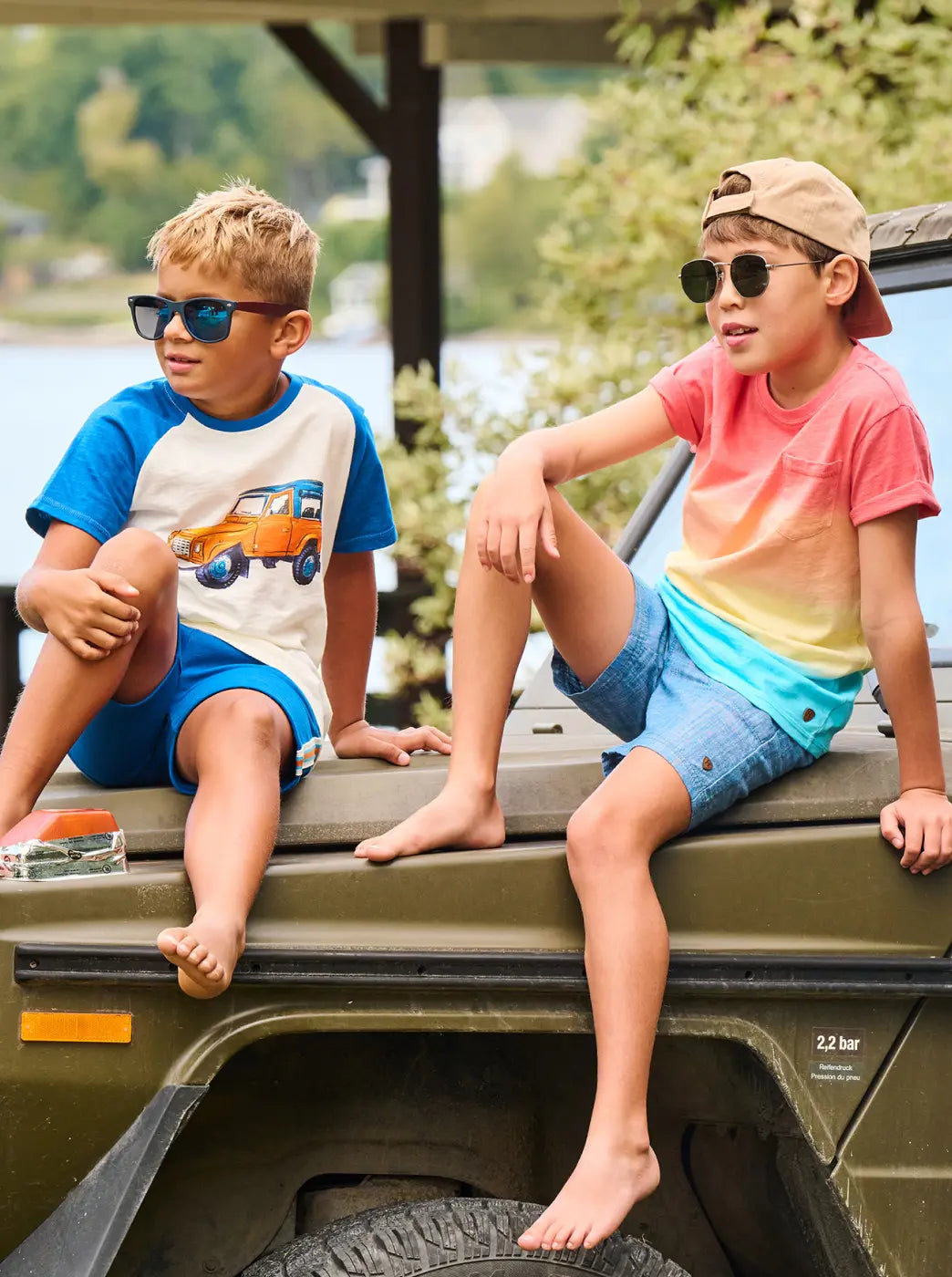 Shop boys' clothing collection. Showing two boys in colorful summer outfits and sunglasses sit on a vehicle outdoors.