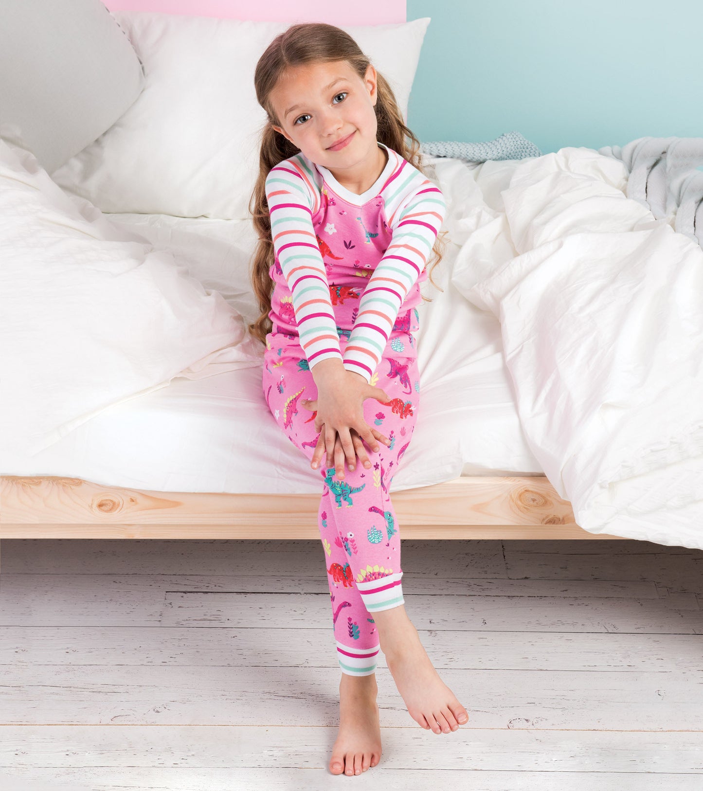 Pink cotton pajama set featuring colorful dinosaur prints and striped sleeves, worn by a girl sitting on a bed.