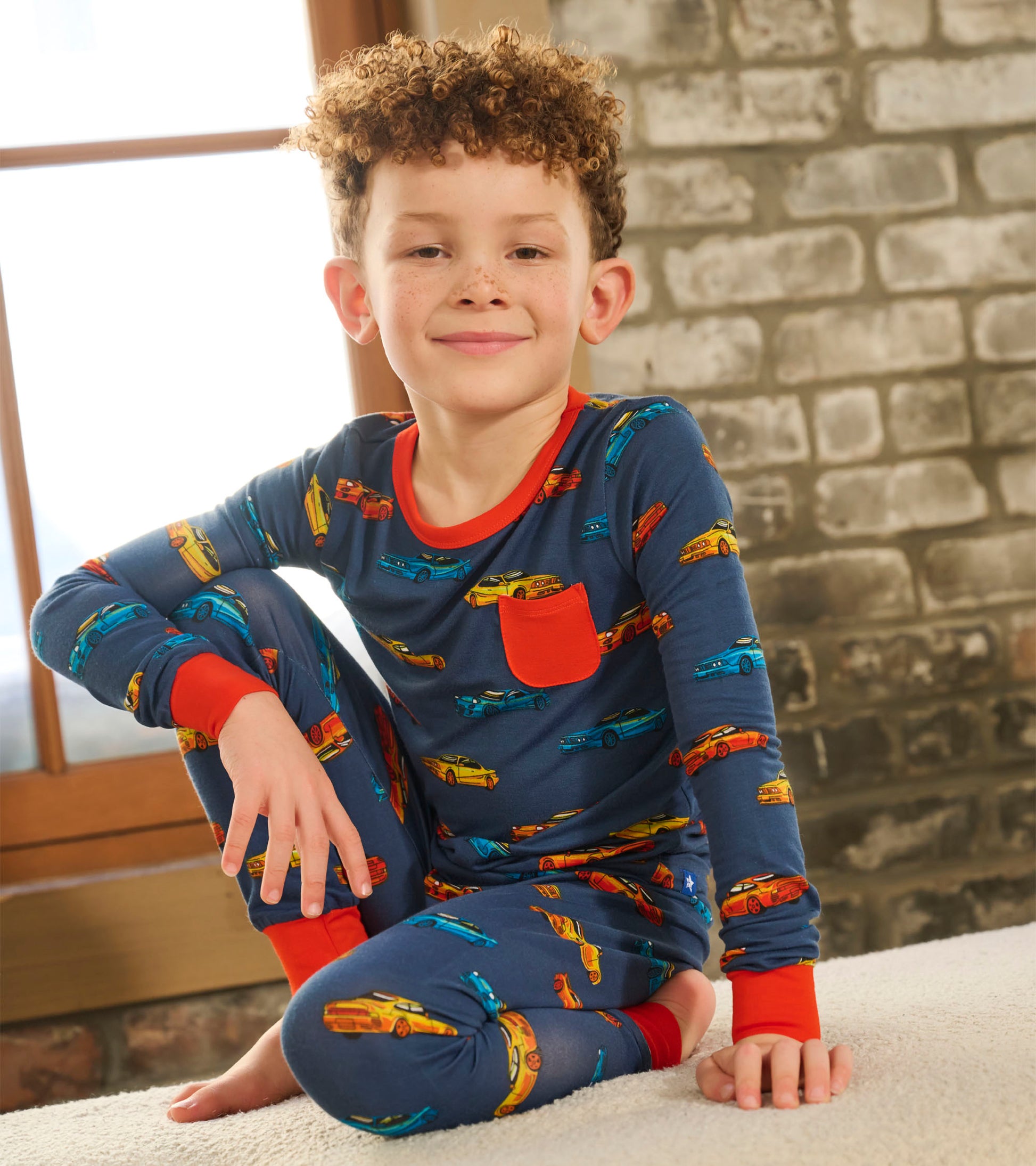 Boy wearing navy long-sleeve pajama set with colorful sports car print and red trim, seated cross-legged