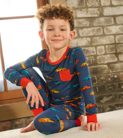Boy wearing navy long-sleeve pajama set with colorful sports car print and red trim, seated cross-legged