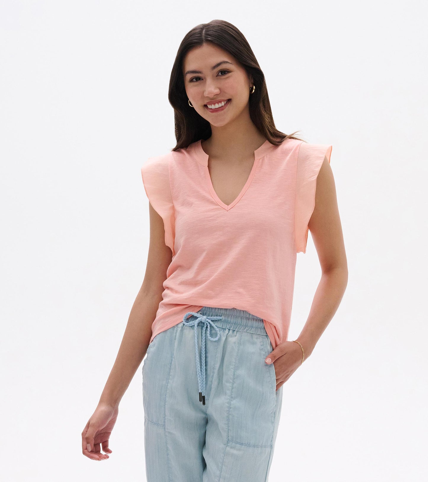Woman wearing a blush pink sleeveless V-neck tank top with flutter sleeves and light blue pants.