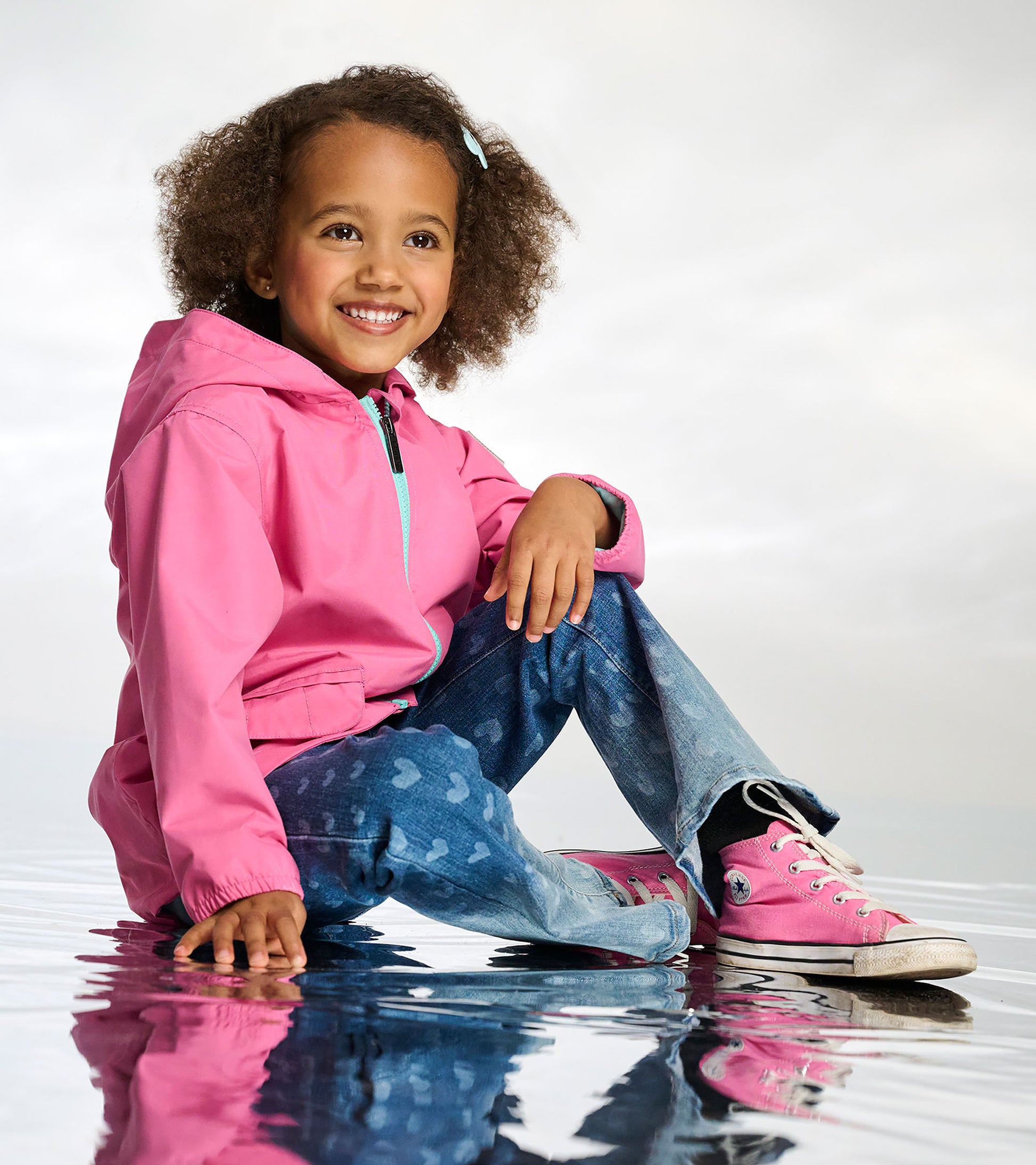 Girl wearing a pink hooded rain jacket with turquoise zipper and blue butterfly-print pants, seated front view