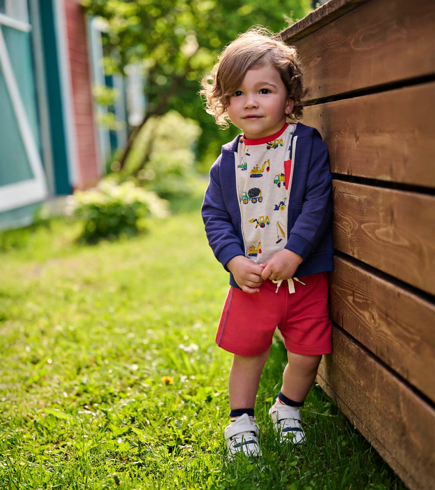 Toddler boy wearing red French terry shorts with navy trim, standing outdoors by a wooden fence