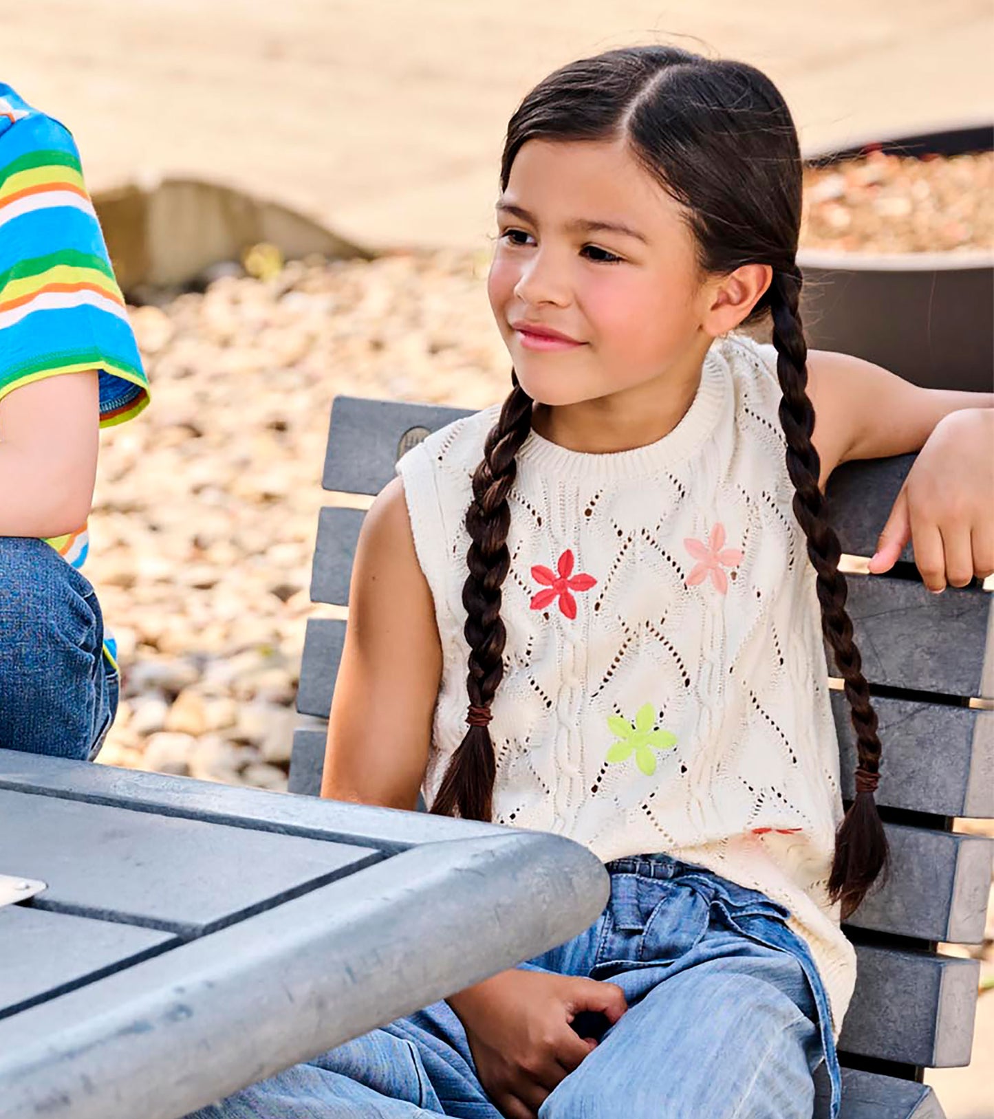 Girl wearing a cream sleeveless knit top with openwork pattern and colorful flower appliqués, seated outdoors