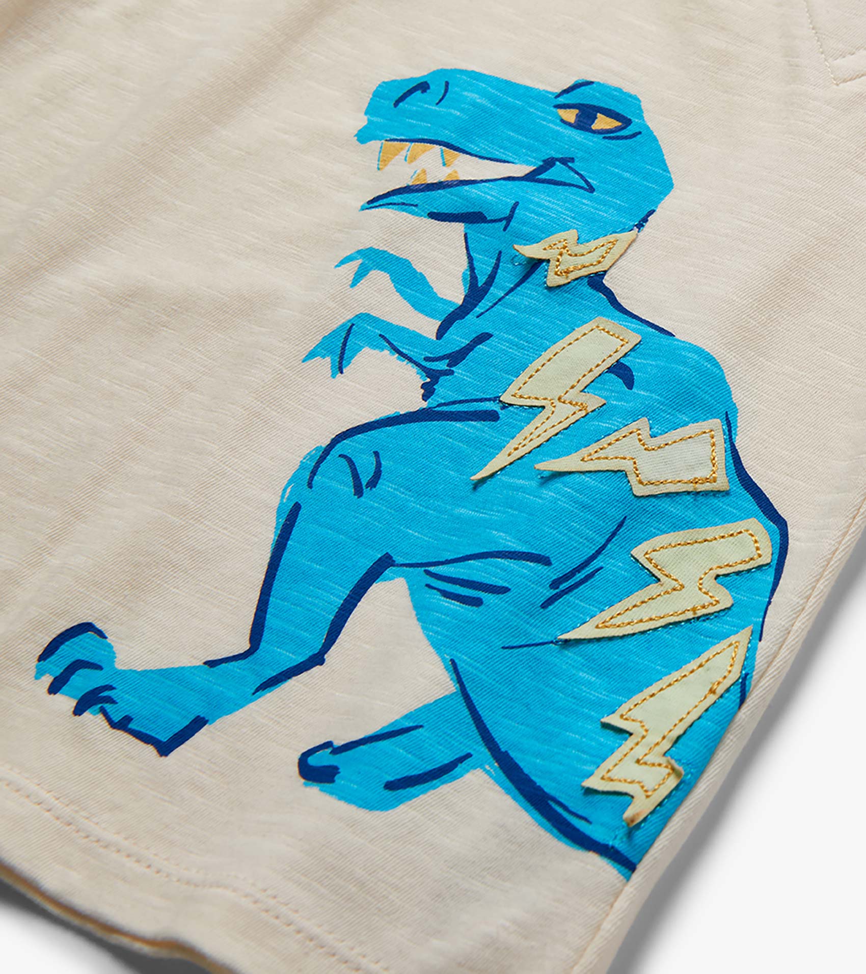 Close-up of beige shorts with a blue dinosaur graphic and lightning bolt patches on the leg.